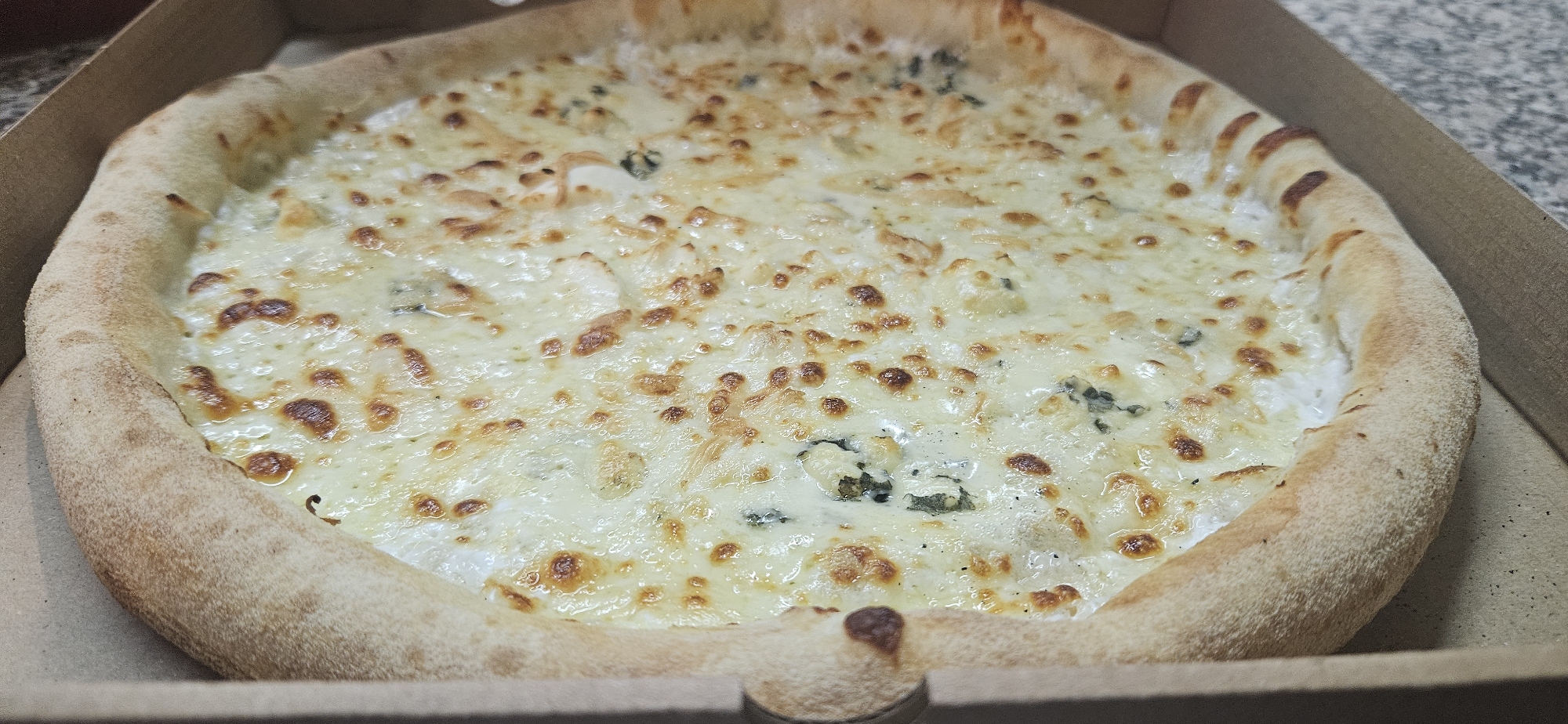 The image shows a pizza inside a cardboard box. The pizza is covered in a white sauce and melted cheese, with occasional green herbs or vegetables visible under the cheese. The crust is thick and puffed up. The image appears to be taken indoors, with the pizza resting on a countertop. The location is stated to be in the city of Sofia, Bulgaria. There are no people present in the image.