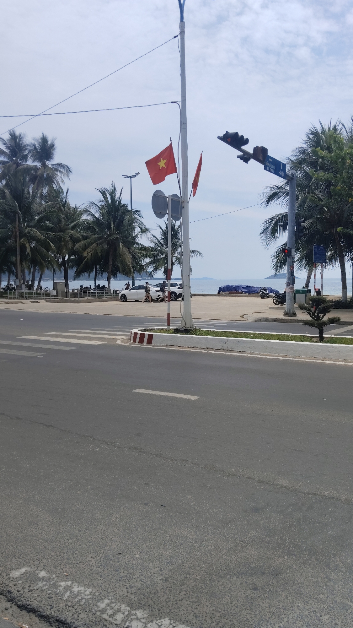 The scene is on a street in Bac Nha Trang Ward, Vietnam. In the background, palm trees line a coastal area with people near the beach. In the middle ground, there is a crosswalk near a traffic light, which is red. A white car is parked near the sidewalk with people nearby. A motorcycle is parked further down the road. The foreground is an asphalt road with a white stripe painted on it. A Vietnamese flag is attached to a utility pole near the traffic light.