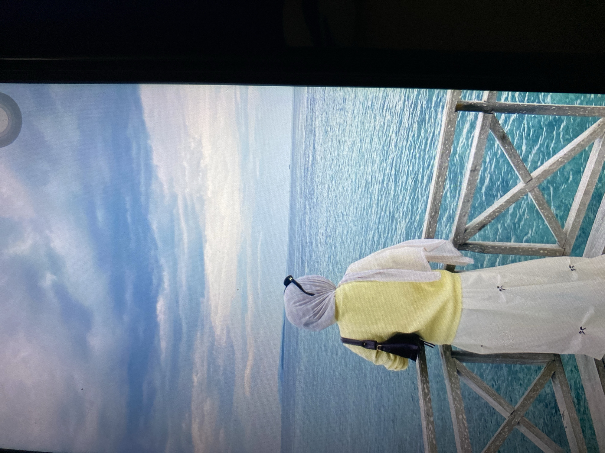 A lone figure, wearing a light yellow long-sleeved shirt, a white hijab, and a long white skirt, stands on a wooden boardwalk overlooking a turquoise ocean under a cloudy blue sky. The person is seen from behind, with a dark handbag slung over their left shoulder and sunglasses perched on top of their hijab. The boardwalk has weathered wooden railings. In the distance, a faint landmass is visible on the horizon, and a small ship is present further out at sea. The scene appears to be a scenic overlook or pier. The image quality is slightly degraded, possibly due to screen capture. No specific location in Ta', Indonesia is identifiable from the visual information presented.