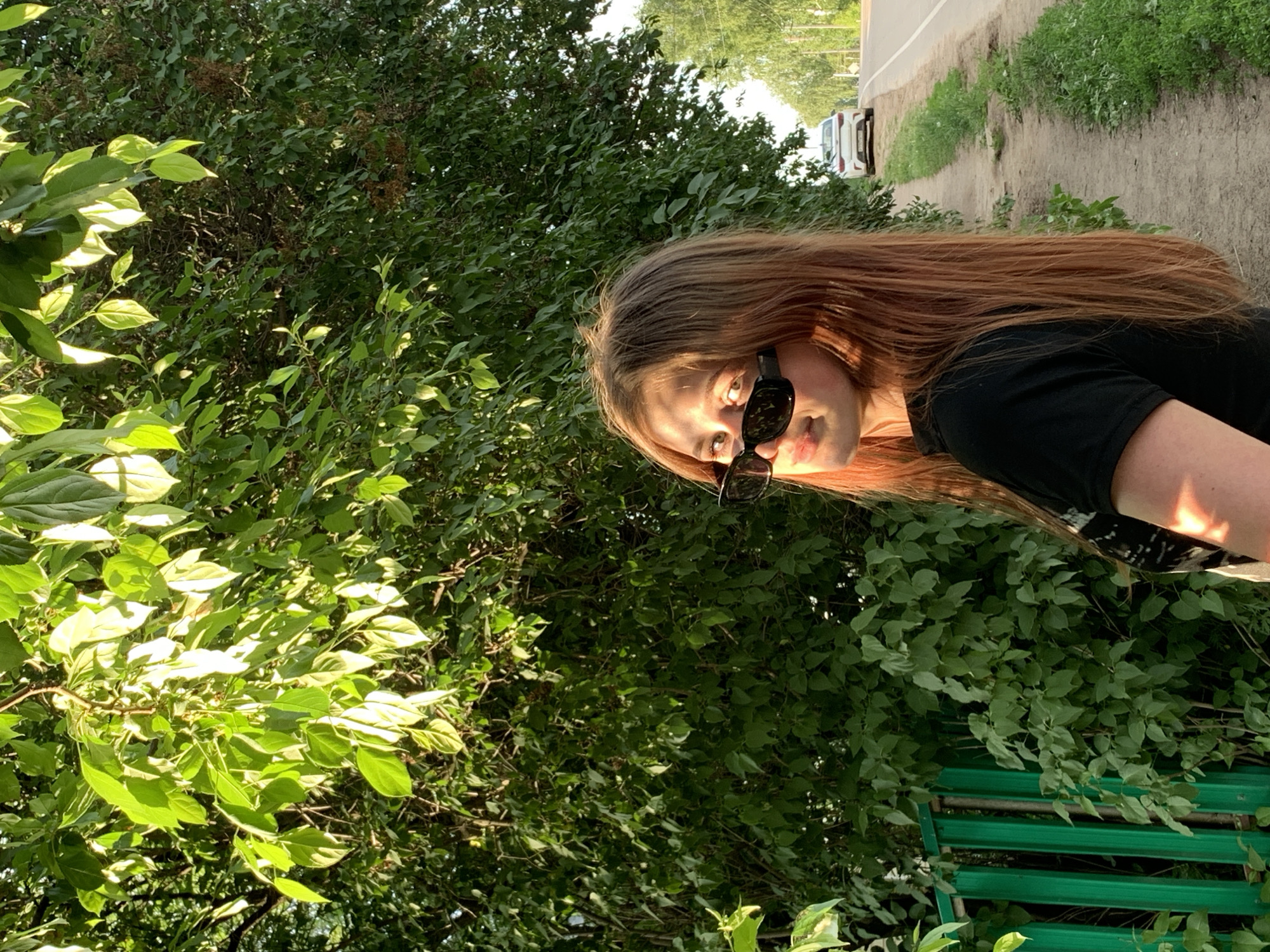 An outdoor, eye-level shot captures a young person posing in bright daylight, alongside a roadside surrounded by lush greenery. The image is oriented vertically (originally rotated 90 degrees counter-clockwise).
The main subject, a person with long, reddish-brown hair, is positioned on the right side of the frame. They are wearing dark sunglasses and a black short-sleeved top. Their head is slightly tilted, and they are looking directly at the camera, with a subtle expression. Bright sunlight highlights their hair and face.
The background is dominated by dense green foliage, appearing as thick bushes or trees, particularly on the left and center. On the upper right, a dirt or gravel path runs parallel to an asphalt road. A white car is visible in the distance on the road. In the bottom right corner, a section of a green, slatted fence is partially visible, also surrounded by green leaves.
The bright, natural lighting suggests it is daytime, likely a sunny day. No discernible text is visible in the image. The setting appears to be a rural or suburban area, possibly a park or roadside in Фоки, Russia.