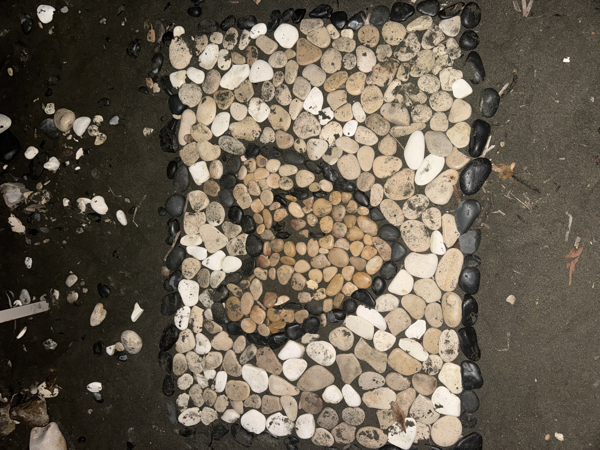 The image depicts a heart shape formed from smooth, rounded pebbles of varying colors, predominantly white, tan, and orange, set against a backdrop of dark sand. The pebble heart is centrally positioned within a larger rectangular arrangement of pebbles, which are themselves embedded in the sand. The surrounding environment appears to be a beach or a sandy area. The lighting suggests it is nighttime. The image does not contain any people or discernible actions. This scene is located in the city of Agios Tychona Community, Cyprus.