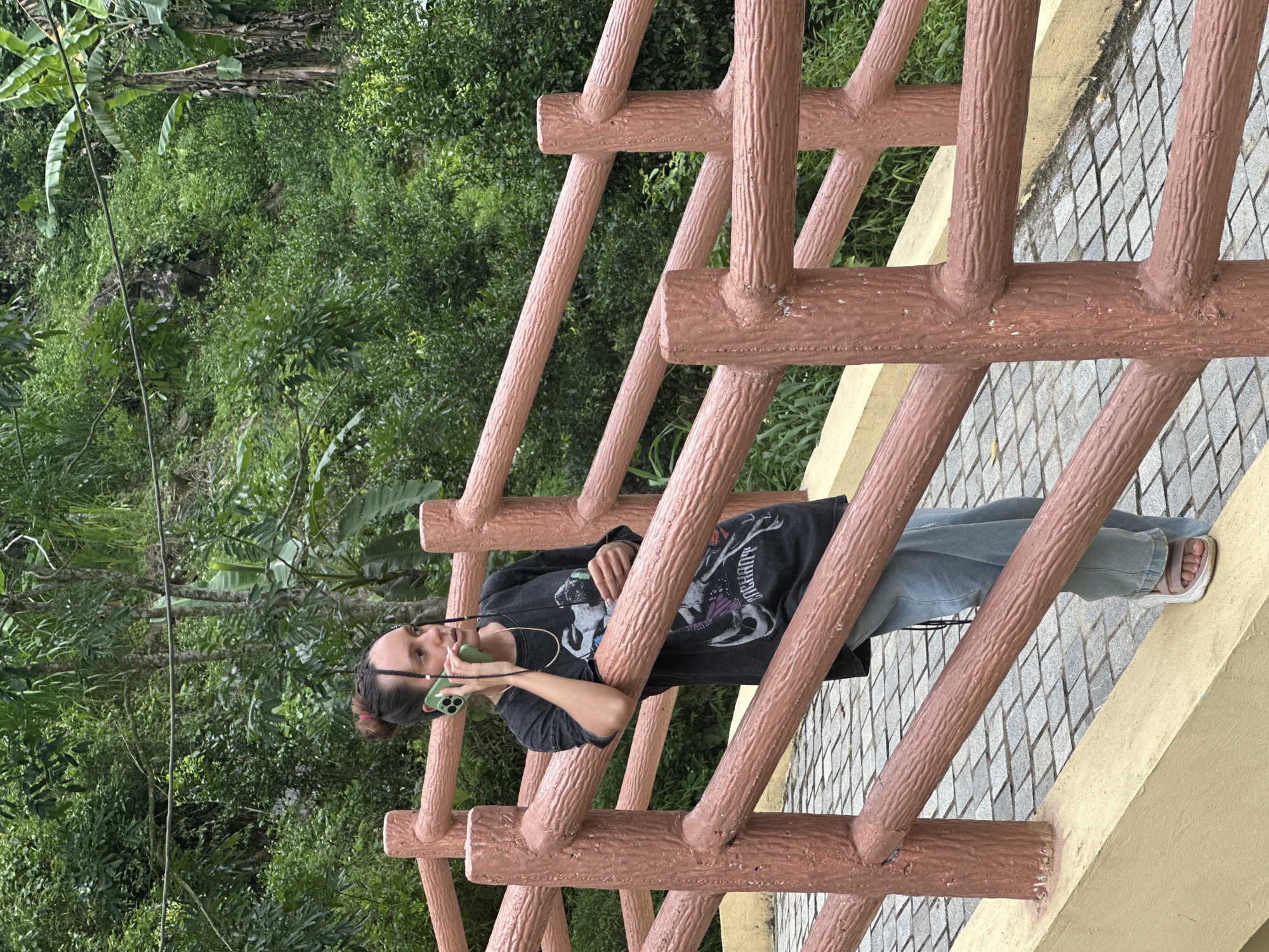 A young woman with dark hair styled in a bun and two braids is standing on a stone pathway and leaning against a wooden railing. She is holding a green smartphone to her ear with her left hand and wearing a dark t-shirt with a graphic print, light blue jeans, and light-colored sandals. The railing has a reddish-brown, wood-like texture. The background consists of lush green foliage and trees, suggesting an outdoor, natural environment. The location appears to be in a tropical or subtropical region, consistent with Pù Luông Commune, Vietnam, known for its natural beauty and mountainous landscape.