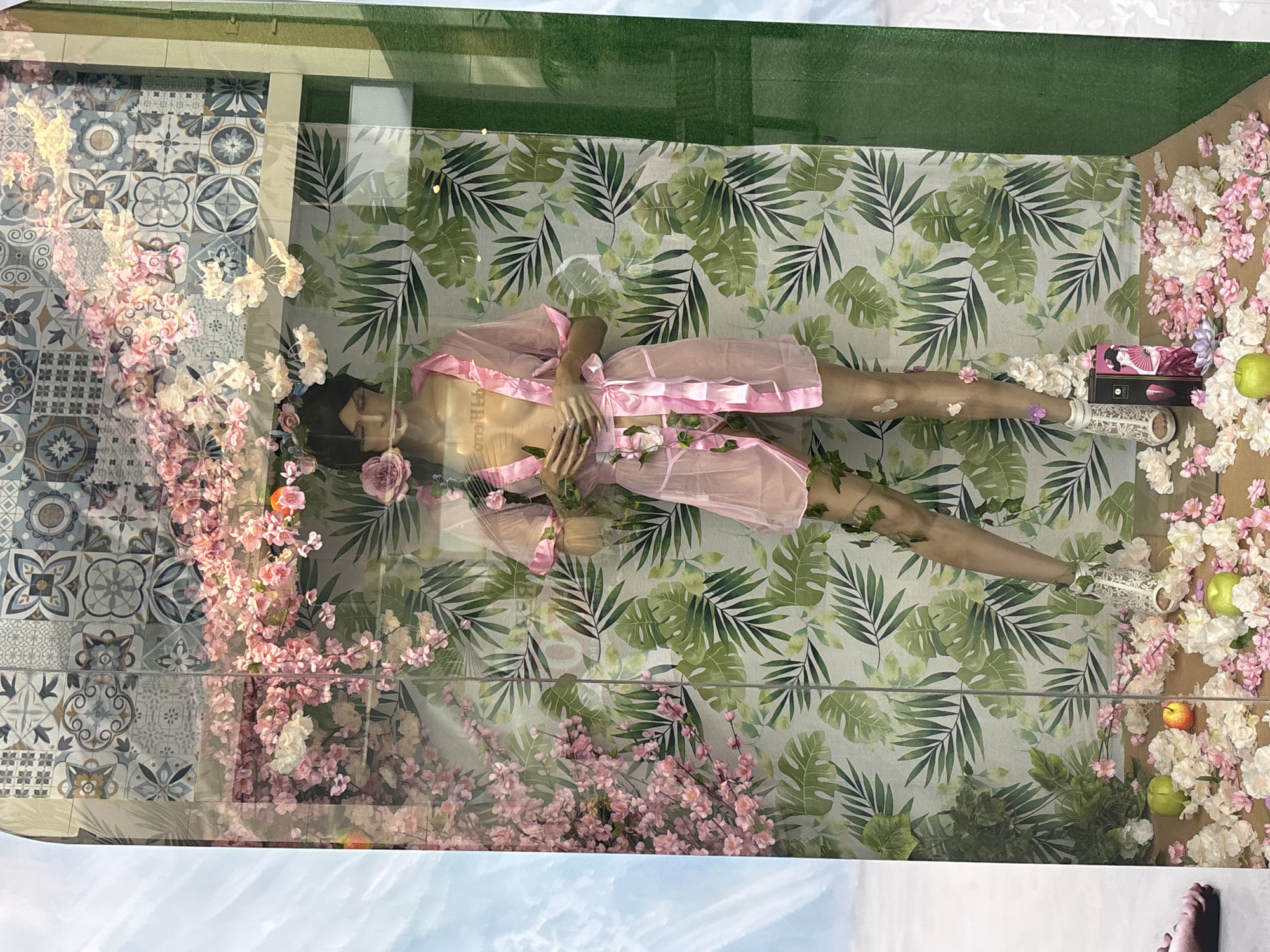 The image displays a mannequin in a store window in Orihuela, Spain. The mannequin is wearing a sheer pink robe with a matching belt, adorned with pink ribbon and artificial flowers. The background of the window features a tiled wall with a repeating pattern of blue and white geometric designs, and a tropical leaf-patterned wallpaper. Pink and white artificial flowers cascade from the top of the window, and vines with artificial leaves are draped around the mannequin. The floor of the display is covered with artificial flowers, two green apples, and one orange apple. A dark-colored box with an image of a geisha on it is placed on the floor near the mannequin's feet. The overall scene is a highly stylized and decorative display, likely for a clothing or lingerie store.