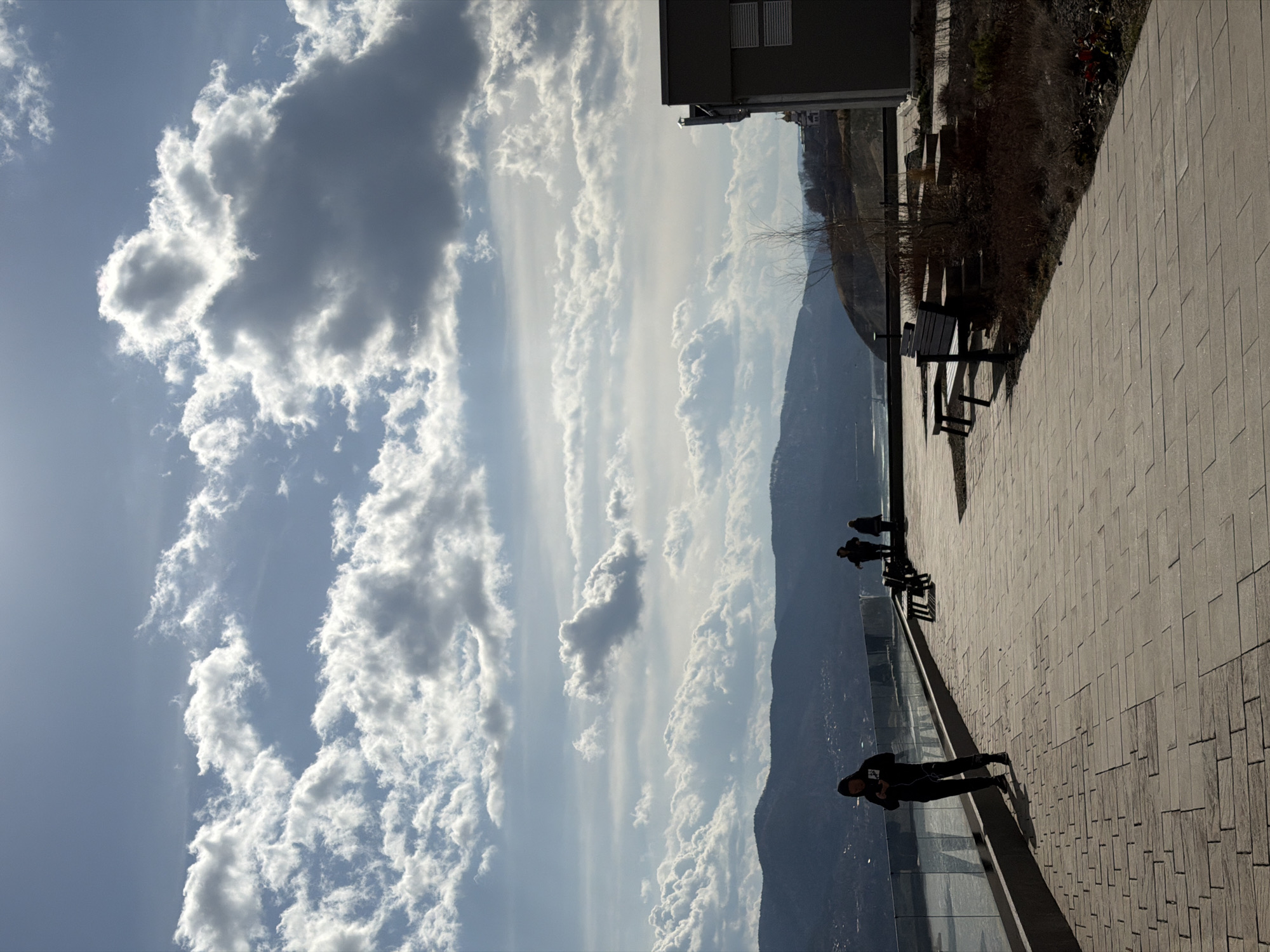 The image shows a promenade in Krasnoyarsk, Russia, under a partially cloudy sky. The view overlooks a mountain range and a city visible in the valley below. A paved walkway, with park benches, stretches along the edge of the view. Several individuals are present: One person stands near the glass barrier, appearing to look out at the view, and other individuals stand farther down the walkway. A building is on the upper right.