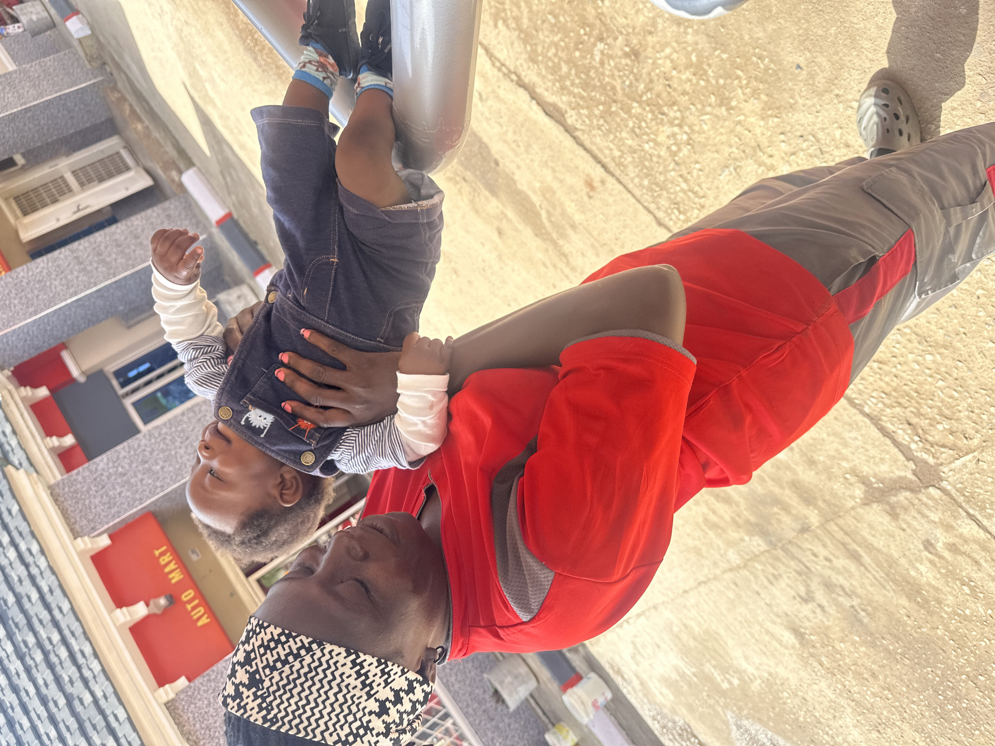 An adult, wearing a red shirt and a black and white patterned headscarf, holds a baby wearing a denim overall and a striped long-sleeved shirt. The adult's eyes are closed, and they appear to be looking down at the baby. The baby is looking upwards and to the right. In the background, there is a building with a red awning that says "AUTO MART" in yellow lettering, indicating the location is a business in Gura Top, Nigeria. The ground is a light-colored, textured surface, possibly concrete or asphalt, with some shadows cast by the building and the people. The overall lighting suggests it is daytime, with sunlight illuminating the scene.