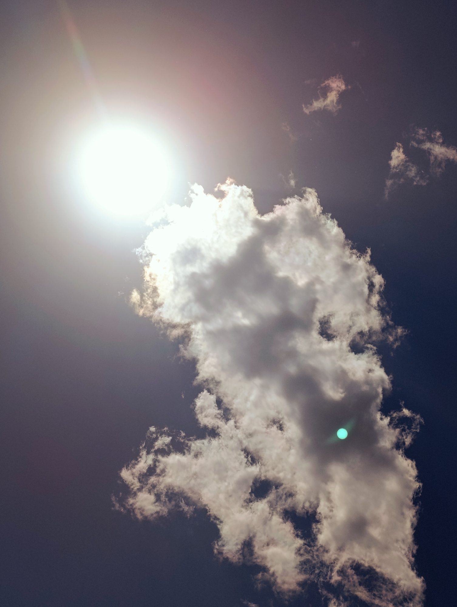 A vertical, wide shot captures a portion of the sky over Naryn City, Kyrgyzstan. The sun, a bright white orb with a faint pinkish-red halo, dominates the upper left quadrant. Rays of light emanate from it, creating a hazy effect across the upper half of the frame. A large, dense, vertically oriented cloud, tinged with gray and white, stretches from the center towards the bottom of the image. Within this cloud, a small, bright turquoise-green circular lens flare is visible. The sky transitions from a bright, hazy gray near the sun to a deep, dark blue towards the right and bottom. Scattered across the sky are smaller, wispy white clouds.