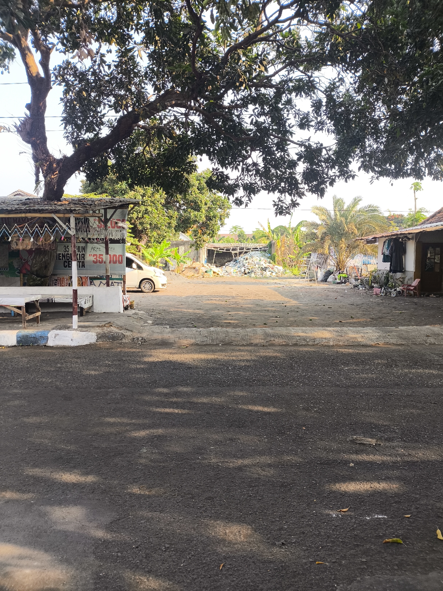The image depicts an outdoor scene in the city of Citrodiwangsan, Indonesia, likely in a rural or semi-urban area. In the foreground, a paved road with asphalt texture is visible, with dappled sunlight creating a pattern of light and shadow. To the left, a small roadside stall with a corrugated metal roof stands. A white car is parked in front of the stall, which displays various signs, one of which shows the price "35.100". A wooden table and chairs are also present near the stall.
Behind the stall, the ground is a mixture of packed dirt and gravel. Further back, a large pile of discarded materials, likely plastic waste and debris, is visible, suggesting a waste collection or informal recycling point. Several structures with tin roofs are scattered throughout the area. To the right, a small house or building is situated with clothes hanging outside. A palm tree stands prominently in the mid-ground, surrounded by lush green vegetation, including banana trees. A large, mature tree with dense foliage dominates the upper portion of the image, casting a shadow over the scene. The sky is bright and appears clear, indicating a sunny day. No people are actively depicted in the image. The overall impression is of a functional but somewhat unkempt area, typical of many developing regions.