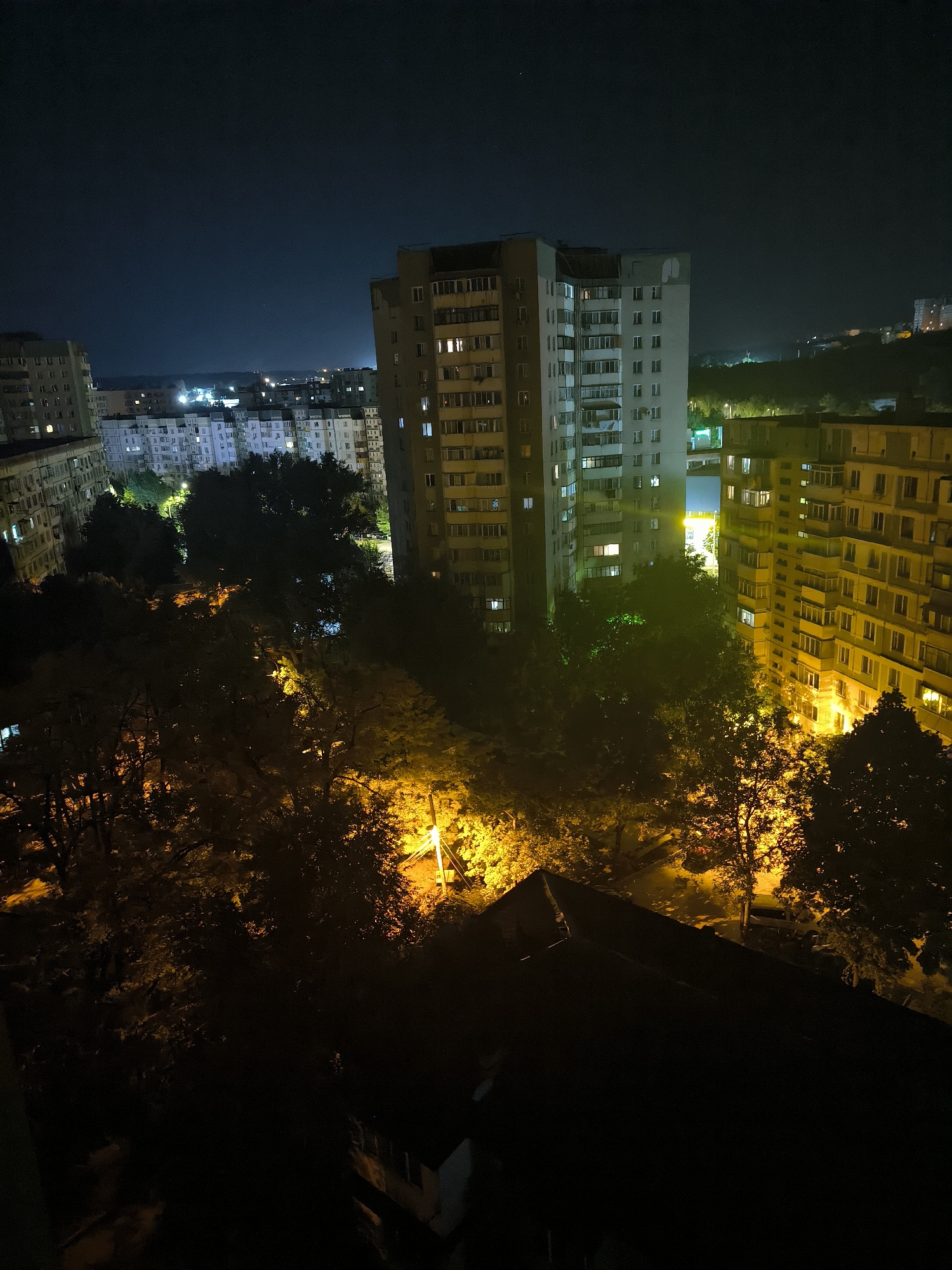 This is a night-time aerial photograph of a residential area in Chișinău, Moldova. The scene is dominated by several multi-story apartment buildings, typical of Soviet-era architecture, some of which are illuminated from within, suggesting occupancy. Lush, dark trees with some illuminated foliage are interspersed between the buildings, casting shadows. Streetlights emit a warm, yellow glow, illuminating parts of the foliage and what appears to be a street or pathway below. The sky is dark, with a slight gradient of lighter blues and grays towards the horizon, indicating the presence of distant city lights. In the foreground, the dark silhouette of a building's roof is visible, suggesting the photograph was taken from a higher vantage point. The overall impression is that of a quiet, urban residential neighborhood at night.