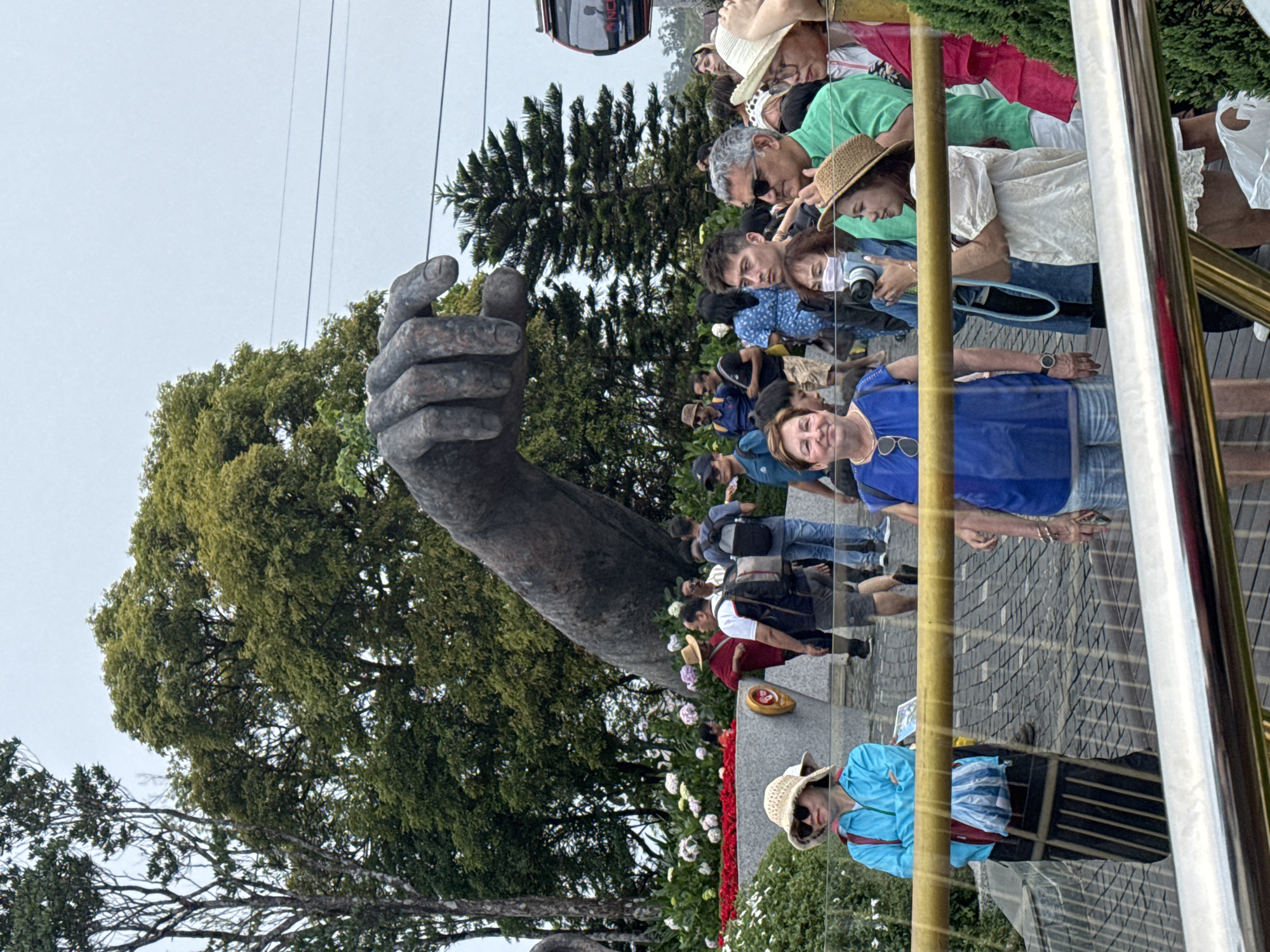 This image depicts a lively outdoor scene at the Golden Bridge in Ba Na Hills, Đà Nẵng, Vietnam. The prominent subject is the massive, weathered stone-like hand sculpture, a distinctive feature of the bridge, which rises from a bed of greenery on the left side of the frame.
A diverse crowd of people, appearing to be tourists, fills the right side of the image. They are casually dressed, with many wearing hats and sunglasses, indicating a bright, daytime setting. Several individuals are seen observing their surroundings, some holding cameras or mobile phones, suggesting they are capturing memories or navigating the site. A woman in a blue top and straw hat is visible in the foreground, partially reflected by a clear railing that defines the visitor pathway.
Lush green trees and dense foliage create a verdant backdrop throughout the scene, contrasting with the paved ground underfoot. Above the landscape, a red cable car cabin, bearing the text "BA NA," can be seen against a bright, overcast sky, further confirming the location as part of the Ba Na Hills resort complex. The atmosphere appears relaxed, with visitors enjoying the scenic landmark. Flower beds with red and white/purple blossoms are visible along the path.