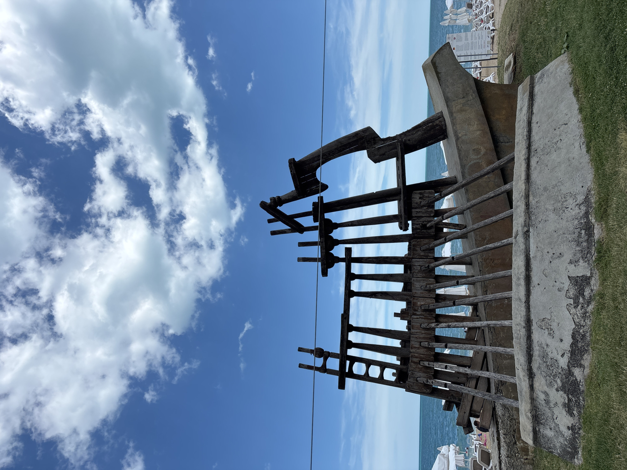 The image depicts a wooden sculpture of a ship's hull and rigging, resembling a partially dismantled vessel. The sculpture is situated outdoors, with the sea and a blue sky with white clouds forming the backdrop. In the distance, a beach with rows of white lounge chairs and umbrellas is visible, suggesting a coastal resort or public beach area. A single thin wire stretches horizontally across the frame, passing in front of the sculpture. The foreground shows a grassy area with a concrete structure, possibly part of the sculpture's base or an older foundation. No people are clearly visible in the immediate vicinity of the sculpture, though a few figures are faintly discernible on the distant beach. The overall scene is that of an art installation at a seaside location. This sculpture is located on the Varna Sea Garden promenade in the city of Varna, Bulgaria, specifically near the entrance to the Sea Garden from the city center.