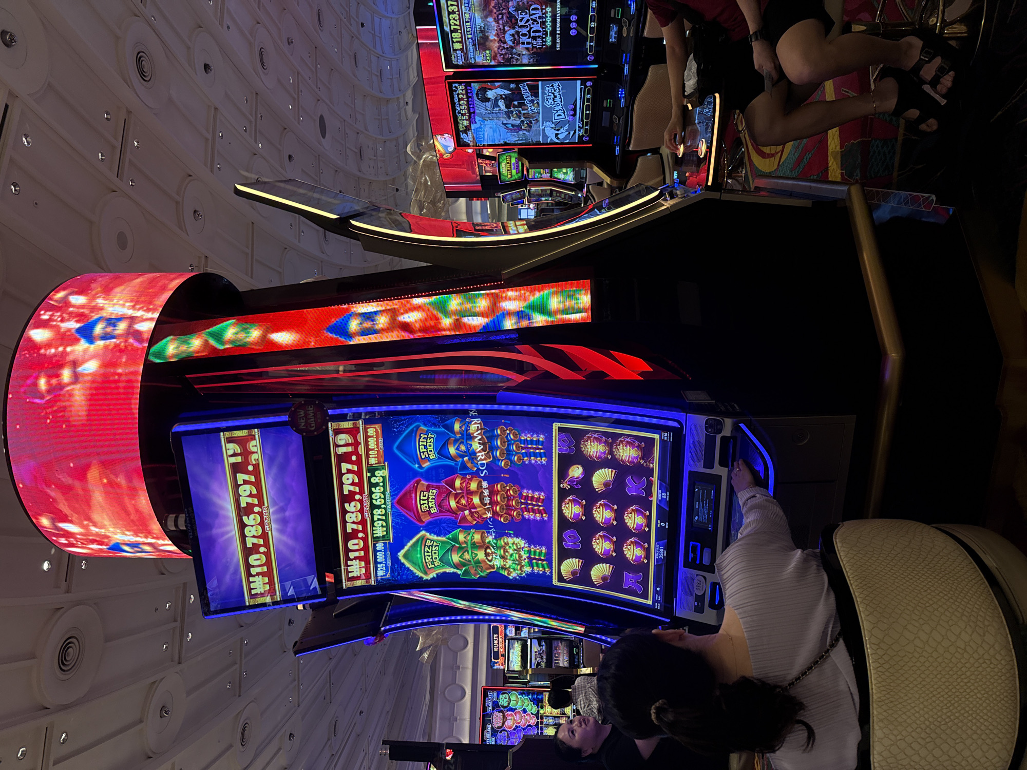 The image depicts the interior of a casino, likely located in Incheon, South Korea. In the foreground, a woman with dark hair tied back is seated in a textured beige chair, her back to the camera, facing a slot machine. She is wearing a light purple pleated top and a dark crossbody bag. Her right hand is extended towards the slot machine's control panel. To her right, another person, wearing a red shirt and black shorts, is seated and is also interacting with a slot machine, their right hand is on the controls.
The prominent slot machine in the foreground is large and brightly lit, featuring a vibrant display with cartoon-like rocket symbols labeled "PRIZE BOOST," "BIG BANG," and "SPIN BOOST." The screen also shows jackpot amounts, including "₩10,786,797.19" for the "GRAND" prize. Other smaller jackpot amounts such as "₩978,696.88" and "₩10,000.00" are also visible. The machine has a modern design with blue and purple accents and a large, curved digital screen above it displaying flashing red graphics with figures that appear to be warriors.
The background reveals rows of other slot machines, some with screens showing different game graphics and jackpot amounts. The casino environment is characterized by bright, artificial lighting and a high ceiling with circular light fixtures. The floor appears to be carpeted with a patterned design. The overall impression is one of a lively and technologically advanced gaming establishment.