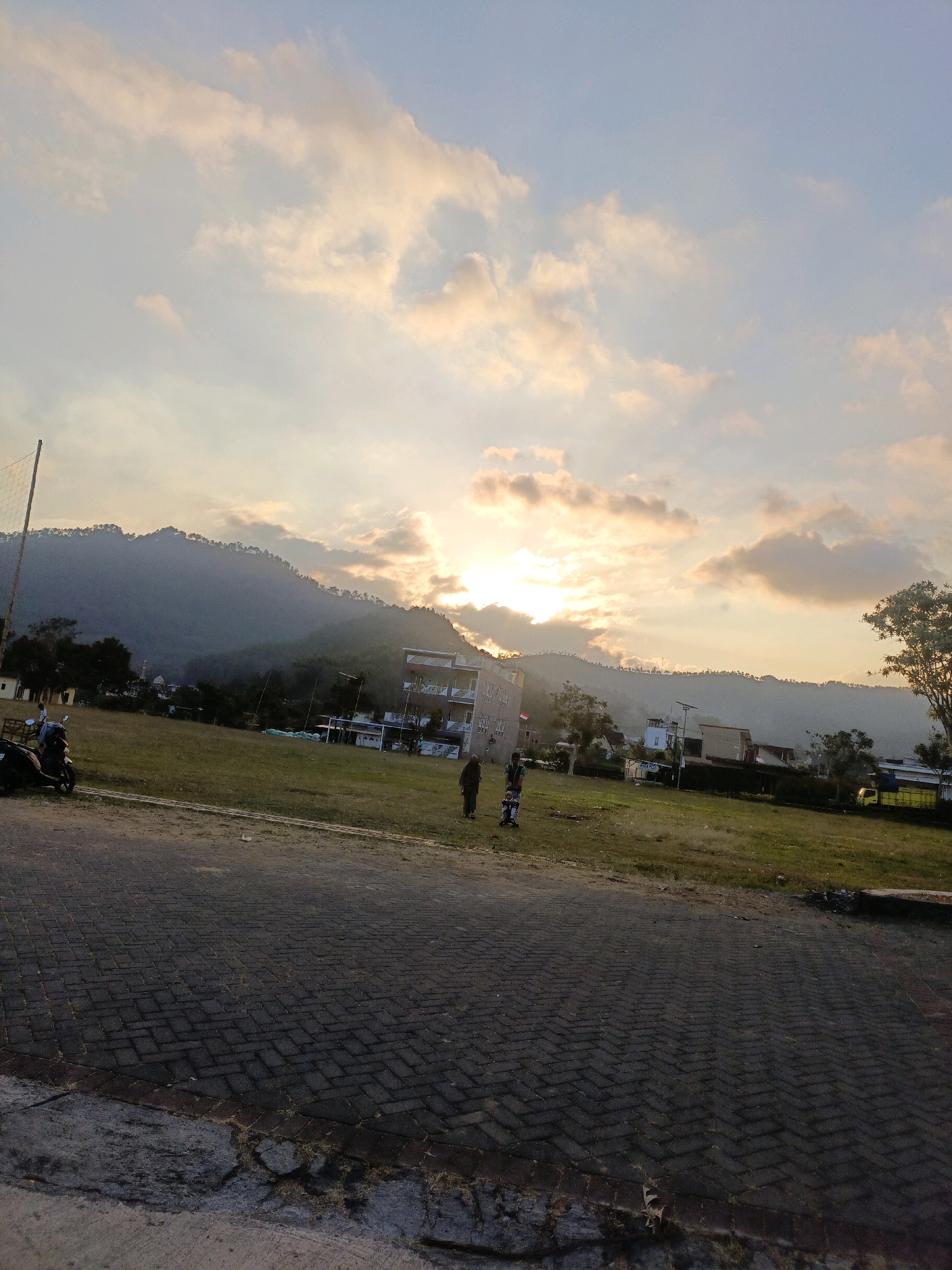 The image depicts a sunset scene at a field, possibly a park or a sports ground, in the city of Batu, Indonesia. The foreground is dominated by a paved area made of dark interlocking bricks, with some patches of concrete and scattered debris. A grassy field stretches beyond the paving, leading to a line of trees and buildings, including a multi-story structure with balconies. In the background, a lush green mountain range rises against the sky. The sky is filled with dramatic clouds, tinged with orange and yellow hues from the setting sun, which is partially obscured by the clouds and the mountain.
Several individuals are present in the scene. On the left side of the field, a person is standing near a parked motorcycle. Closer to the center, two adults and a child in a stroller are walking across the grass. Further to the right, another person is walking on the field. In the distance, a yellow truck is visible. A volleyball net and pole stand on the far left of the field. The overall atmosphere is serene, with the warm light of the sunset casting long shadows and illuminating the landscape. The presence of mountains and the distinctive architecture of the buildings suggest a location in a hilly or mountainous region, consistent with Batu, a city known for its natural beauty and cooler climate.