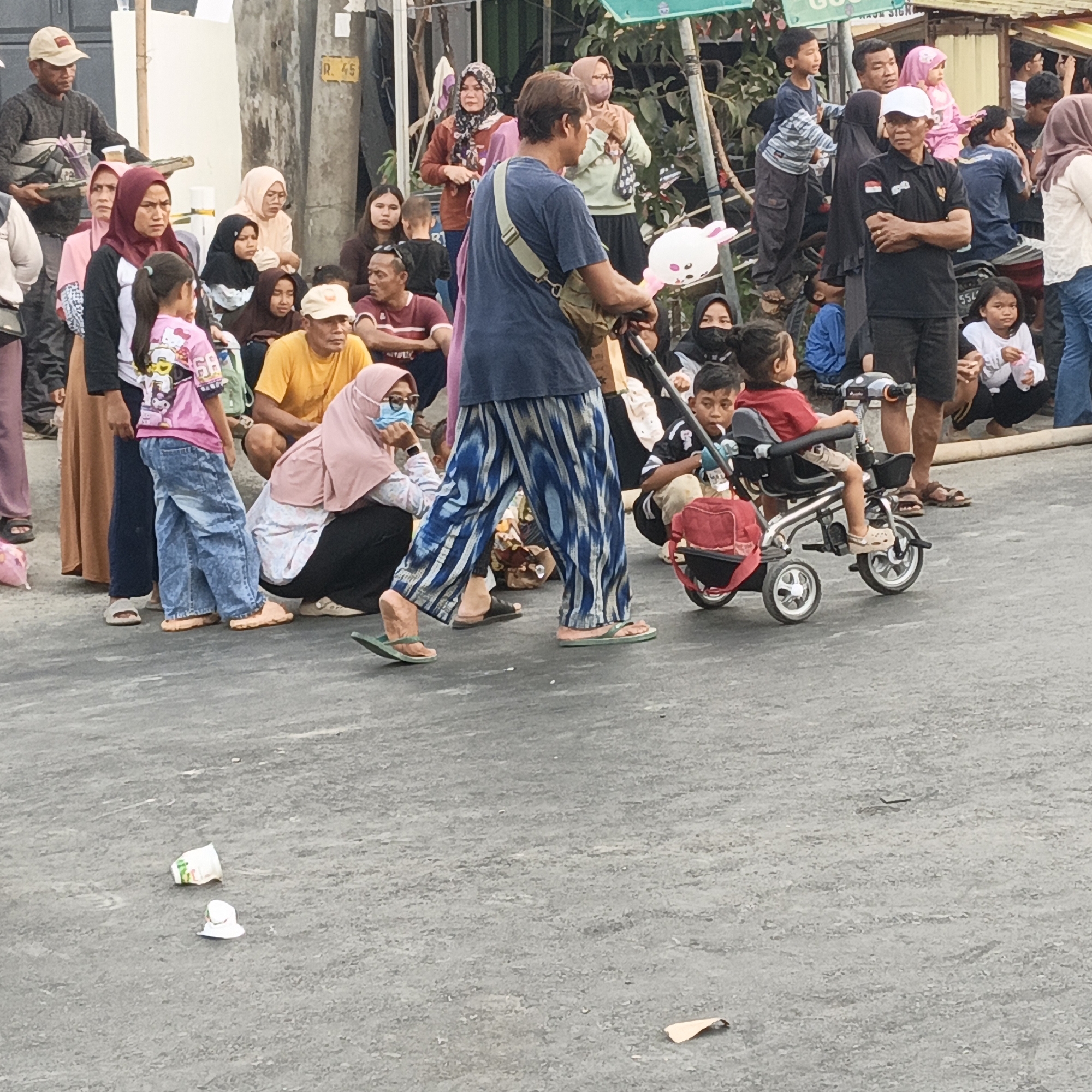 The image depicts a street scene in Randudongkal, Indonesia, with a crowd of people gathered. In the foreground, a man in a blue and white striped shirt and patterned pants pushes a tricycle with a young child seated in it. The child is wearing a red shirt and beige shorts. To the left of the man, a young girl in a pink t-shirt with a cartoon character and blue jeans stands. Further to the left, a woman in a headscarf and brown skirt stands next to another woman in a pink headscarf and brown patterned skirt.
Behind them, a diverse group of people are visible, some sitting and some standing. Many are wearing traditional Indonesian clothing, including headscarves and long skirts. The environment appears to be a street or an open area, possibly a market or a gathering place, with buildings and trees visible in the background. There are some discarded items on the ground, including cups and papers, suggesting a public space that may experience foot traffic. The overall atmosphere seems to be one of a community event or daily life in the town.