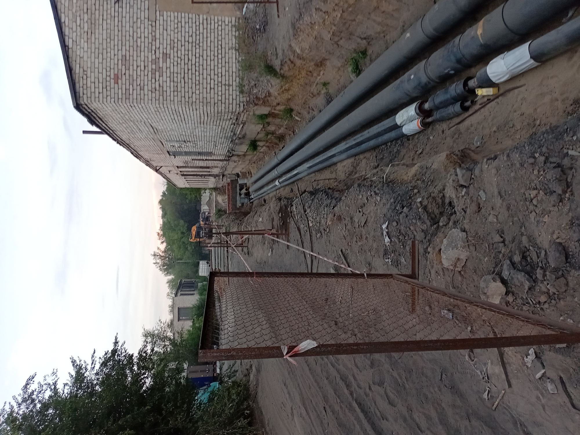 The image depicts an outdoor construction site in Semey, Kazakhstan, characterized by a trench containing large diameter pipes, likely for utilities. The pipes are black and appear to be insulated, with some sections wrapped in white material at the joints. They are laid within a dug trench along the side of a long, single-story building constructed of light-colored bricks. The building has a dark roof and several windows along its facade. To the left of the trench, a sandy path runs parallel to it, bordered by a rusty metal chain-link fence with a post. A red and white ribbon is tied to the fence. Trees and other vegetation are visible in the background, along with a yellow excavator in the distance, indicating ongoing work. The sky is overcast, suggesting a cloudy day. The ground is composed of dirt, rocks, and sand, consistent with a construction area.