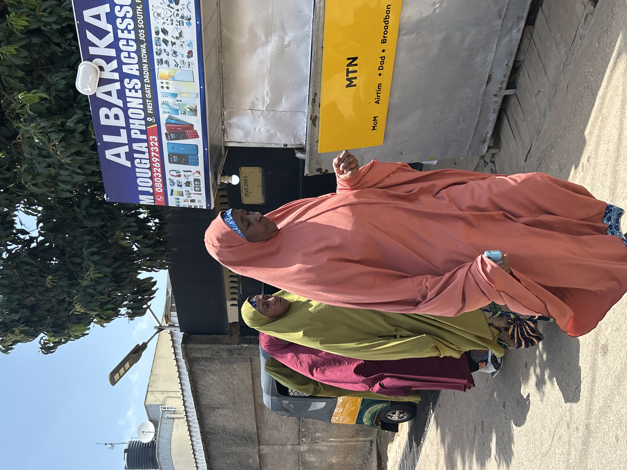 An eye-level outdoor shot captures three women in traditional Islamic attire walking along a paved street in Jos, Nigeria, under a bright sky. The woman in the foreground, wearing a coral-colored hijab and abaya, is captured mid-stride, with her left hand outstretched, holding a light blue object. To her left, two other women are partially visible: one in a dark pink hijab and abaya, and another in a light green hijab and abaya. Behind them, a yellow and green auto-rickshaw is parked on the left side of the street.
Dominating the right side of the frame is a storefront featuring a large, blue sign that reads "ALBARKA M JOUGLA PHONES ACCESSORIES" with a phone number and address. Below this, a white metal structure with a yellow "MTN" logo and service offerings like "MoM, Airtim, Dad, Broadban" is visible. A street lamp is mounted on a pole to the left of the storefront, and a black fence runs along the background. The ground is paved with a rough texture, and shadows are cast by the people and objects, indicating a sunny day. The overall scene depicts a commercial area in Jos.