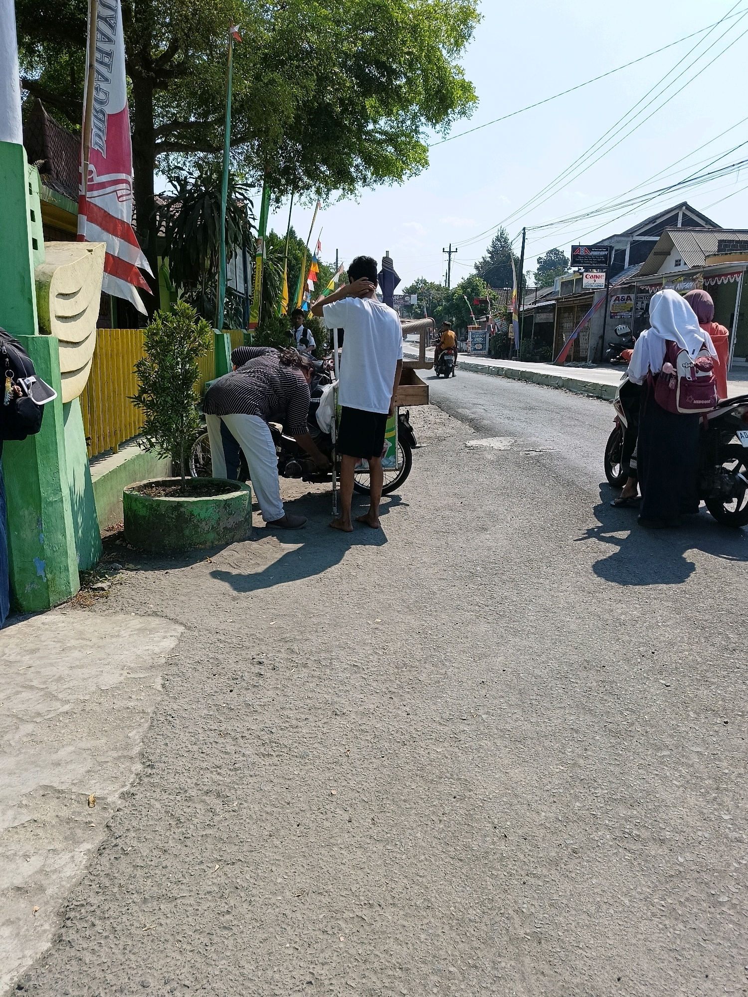 The image depicts a street scene in Barukan, Indonesia, under a bright, clear sky. In the foreground, a person wearing a white t-shirt and black shorts is standing near a motorcycle with a wooden cart attached. Another person, dressed in a striped long-sleeved shirt and white pants, is bending over, appearing to work on the motorcycle. To the right, two individuals, one wearing a white hijab and the other an orange headscarf, are seated on a motorcycle. A third person in a white hijab is standing beside them, holding a red backpack.
The environment includes a paved road with some cracks and patches. On the left side of the road, there's a green and yellow fence and a large tree. Flags are visible on poles near the fence. In the background, buildings line the street, some with signs, including one that appears to be for "Canon." The overall impression is a typical day on a street in an Indonesian town.