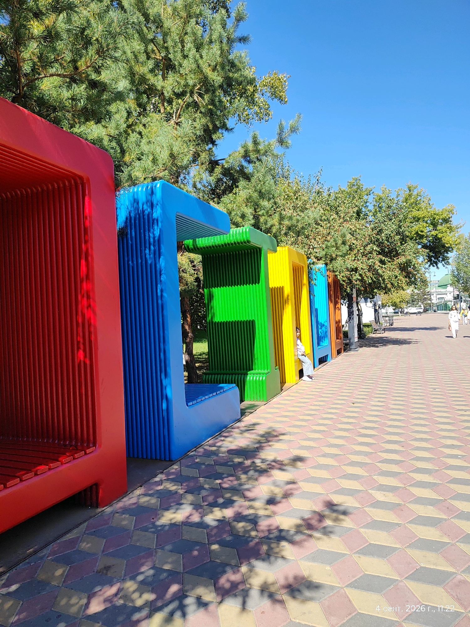 The image shows a row of colorful, oversized letter-shaped seating structures, resembling large, open books, arranged along a patterned walkway. The letters appear to spell out "ETN" in red, blue, green, and yellow, with an orange structure partially visible at the end. A young person is seated inside the yellow structure, looking towards the camera. The walkway is paved with interlocking hexagonal tiles in shades of red, gray, and yellow, creating a 3D optical illusion. Lush green trees line the walkway and extend into the background, with a clear blue sky overhead. In the distance, the facade of a building with a green dome is visible, and several people are walking along the path. The scene is illuminated by bright sunlight, casting dappled shadows from the trees onto the walkway. This is likely a public recreational area, possibly a park or a pedestrian street, in Astana, Kazakhstan, given the presence of specific architectural elements and the Cyrillic date stamp in the lower right corner.