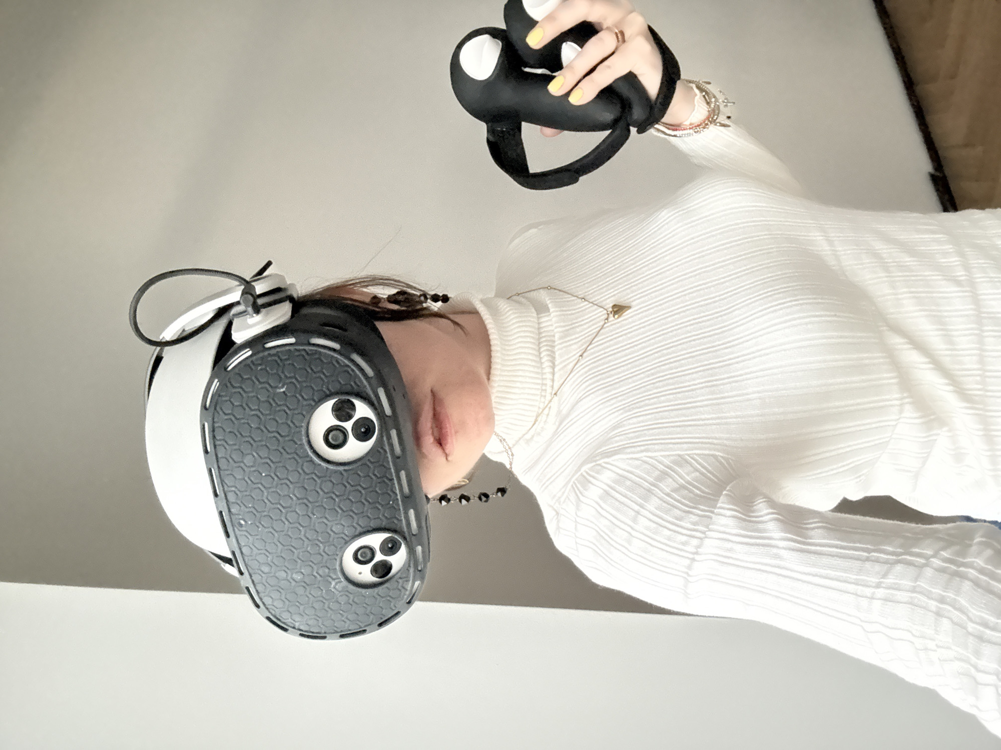 A woman is holding a black and white virtual reality headset and a black controller. She is wearing a white turtleneck sweater, a gold necklace with a heart pendant, and several bracelets on her right wrist. Her fingernails are painted yellow. The background is a plain white surface, possibly a table or wall. The location is not discernible from the given image.