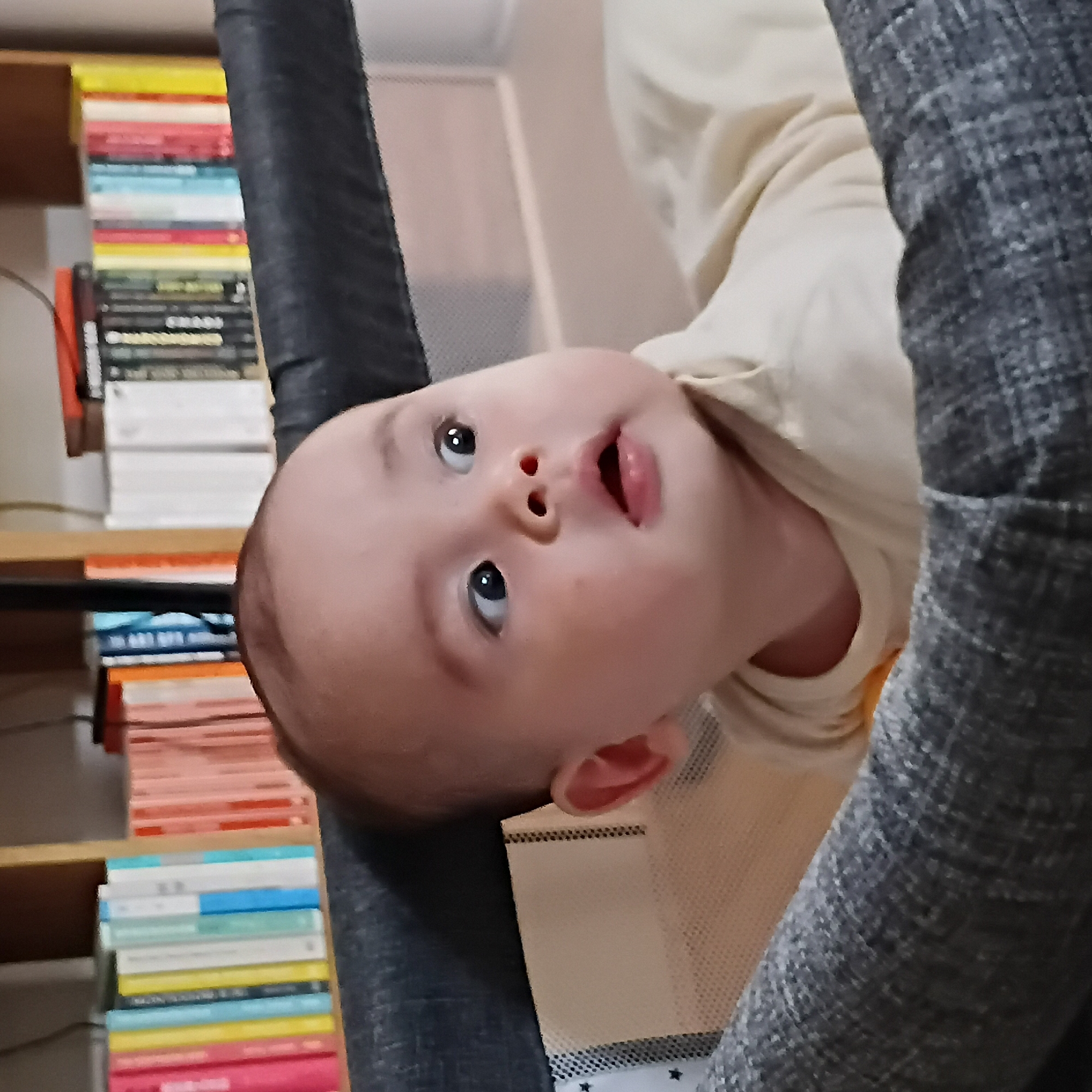 A close-up shot of a baby lying on its back, looking up. The baby has fair skin, dark eyes, and a slightly open mouth. The baby is wearing a light-colored shirt. A bookshelf filled with books is visible in the background. The location is unknown and cannot be determined based on the given information.