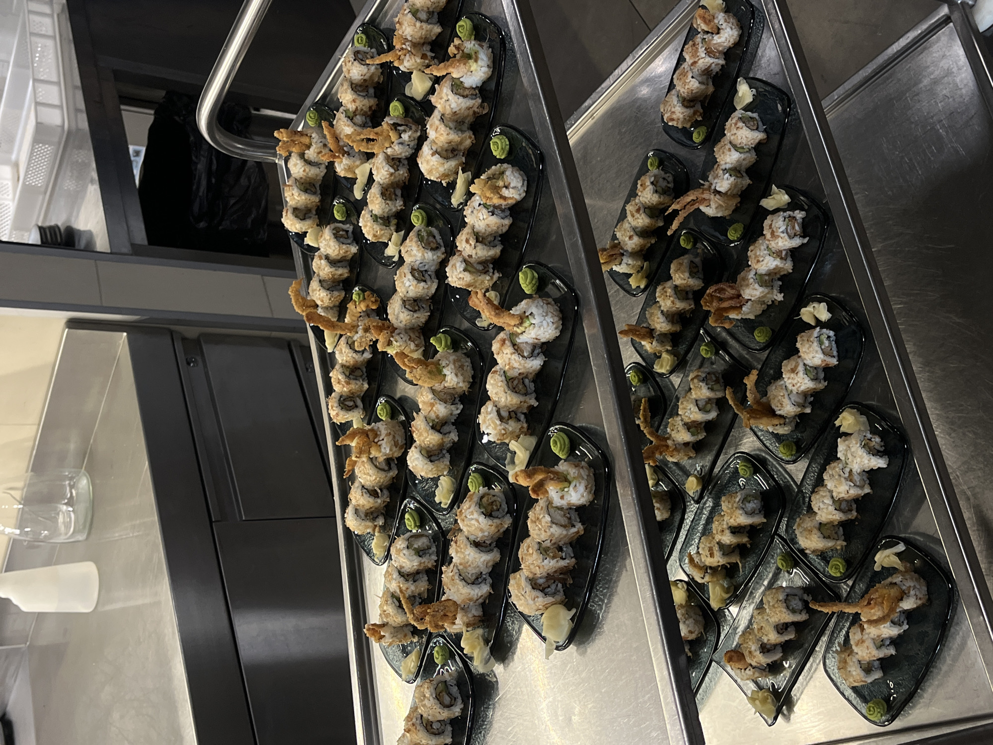 The image shows two stainless steel trays filled with plates of sushi. Each plate holds a row of sushi rolls, garnished with fried topping, ginger, and a dollop of wasabi. The trays appear to be in a commercial kitchen, evidenced by the stainless steel counters, shelving, and equipment visible in the background. The location is not discernible from the image. The presence of sushi suggests a culinary environment, likely preparing for service. There are no people visible.