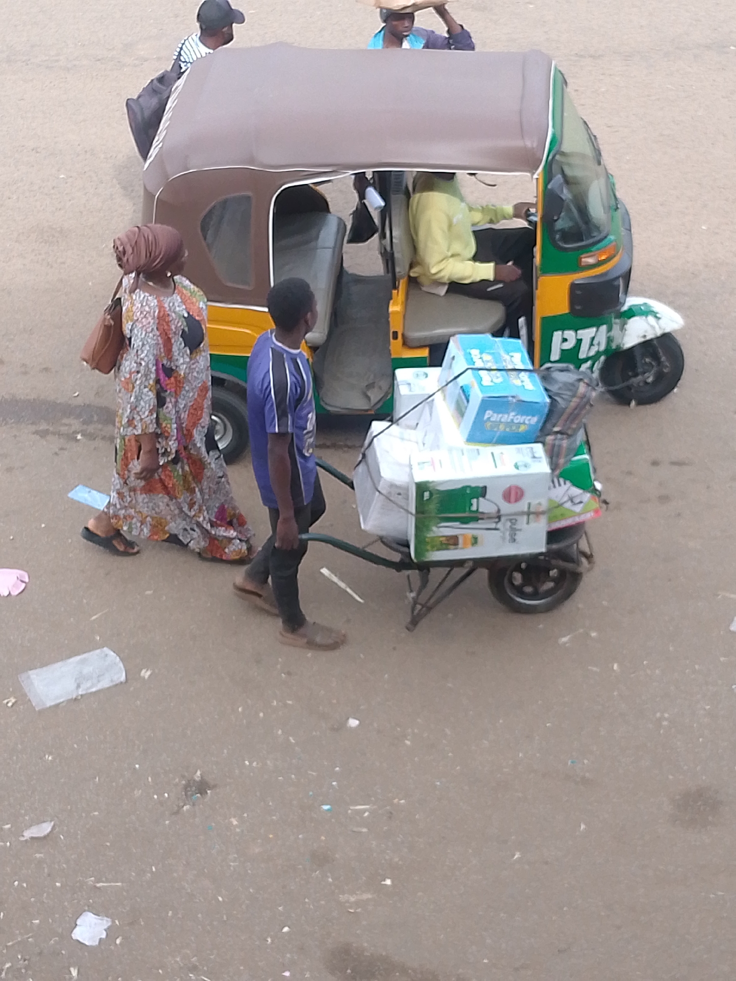 In the city of Bukuru, Nigeria, a scene unfolds on a paved surface, possibly a street or market area. A yellow and green auto-rickshaw, a common mode of transport in the region, is parked. A man, wearing a yellow long-sleeved shirt and dark pants, is seated in the driver's position. A young person in a blue and purple striped shirt and dark pants walks beside the auto-rickshaw, to its left, looking forward. To the left of the auto-rickshaw and slightly behind it, a woman is walking. She wears a long, colorful, patterned dress and a brown headscarf, and carries a brown shoulder bag. Her left foot is visible, wearing a dark sandal.
Attached to the rear of the auto-rickshaw is a small, metal cart. This cart is loaded with several boxes. One prominent box, colored blue and white, displays the text "ParaForce". Another box, white with green accents, shows an image of a sprayer and the word "Pulse". A dark bag is also present on the cart.
In the background, another person wearing a black baseball cap and a striped shirt is partially visible, near the front of the auto-rickshaw. Another individual, wearing a blue garment and carrying something on their head, is also in the background, further back and to the right of the auto-rickshaw. The ground is a muted gray, with some scattered debris, including pieces of white paper and what appears to be a light blue piece of plastic. The overall lighting suggests it is daytime, with the sun casting soft shadows.