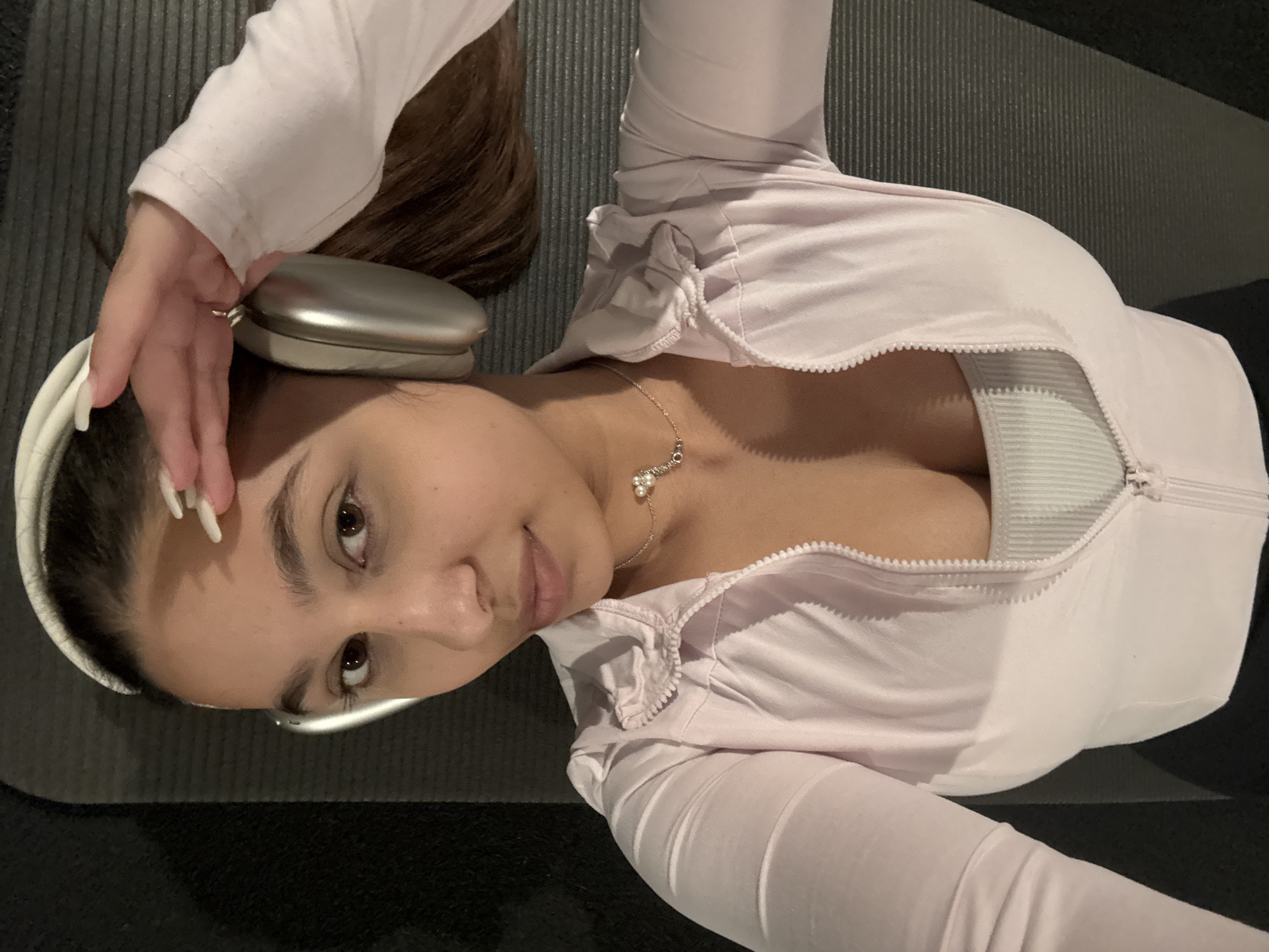 A woman is lying on a black textured surface, possibly a yoga mat or exercise mat. She is wearing a light pink zip-up top with long sleeves, unzipped to reveal what appears to be a white sports bra underneath. A delicate necklace with pearl-like pendants is visible on her chest. She has dark brown hair and is wearing white headphones with silver earcups. Her hand is resting on the earcup closest to the camera, and she has long, light-colored artificial nails. The image is taken from an overhead perspective, looking down at the woman. The setting appears to be indoors. I do not have sufficient information to make a determination regarding the location in the city of Mönchengladbach, Germany.