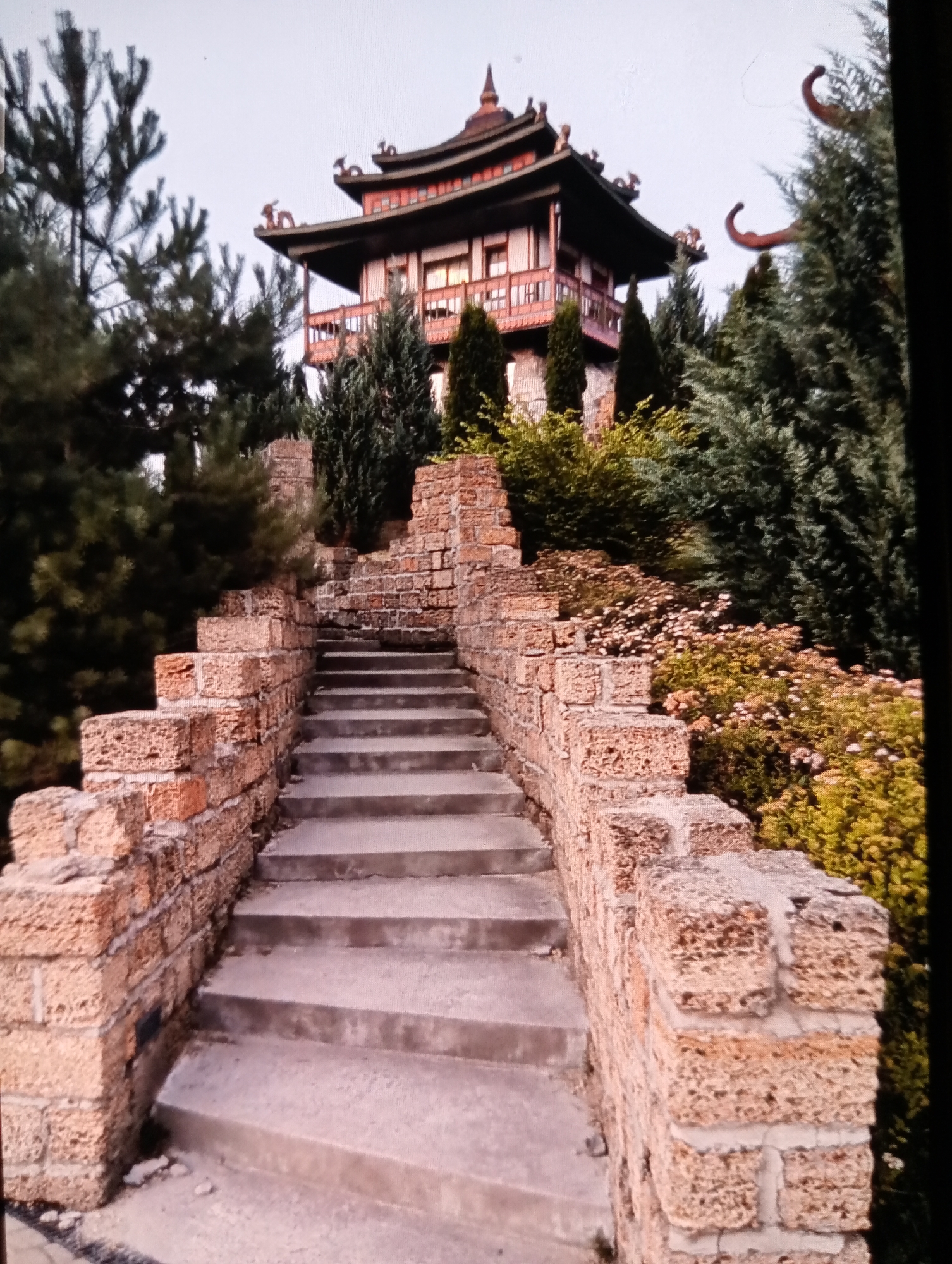 The image shows a stone staircase leading up to a building with a pagoda-style roof. The building is surrounded by trees and bushes. The staircase is made of rough-hewn stones, and the walls flanking the staircase are constructed from similar-looking blocks. The building's architecture is reminiscent of East Asian design, with curved roofs and decorative elements. The surrounding vegetation is lush and appears to be well-maintained. The sky is overcast, suggesting a cloudy day. The scene appears to be an outdoor area, possibly a park or a garden, with the building serving as a focal point. The lighting suggests it is either early morning or late afternoon. There are no people visible in the image. The building and staircase have an aged appearance, suggesting they have been in place for some time. The architectural style and the presence of the pagoda-style roof suggest a deliberate aesthetic choice to evoke a specific cultural theme.