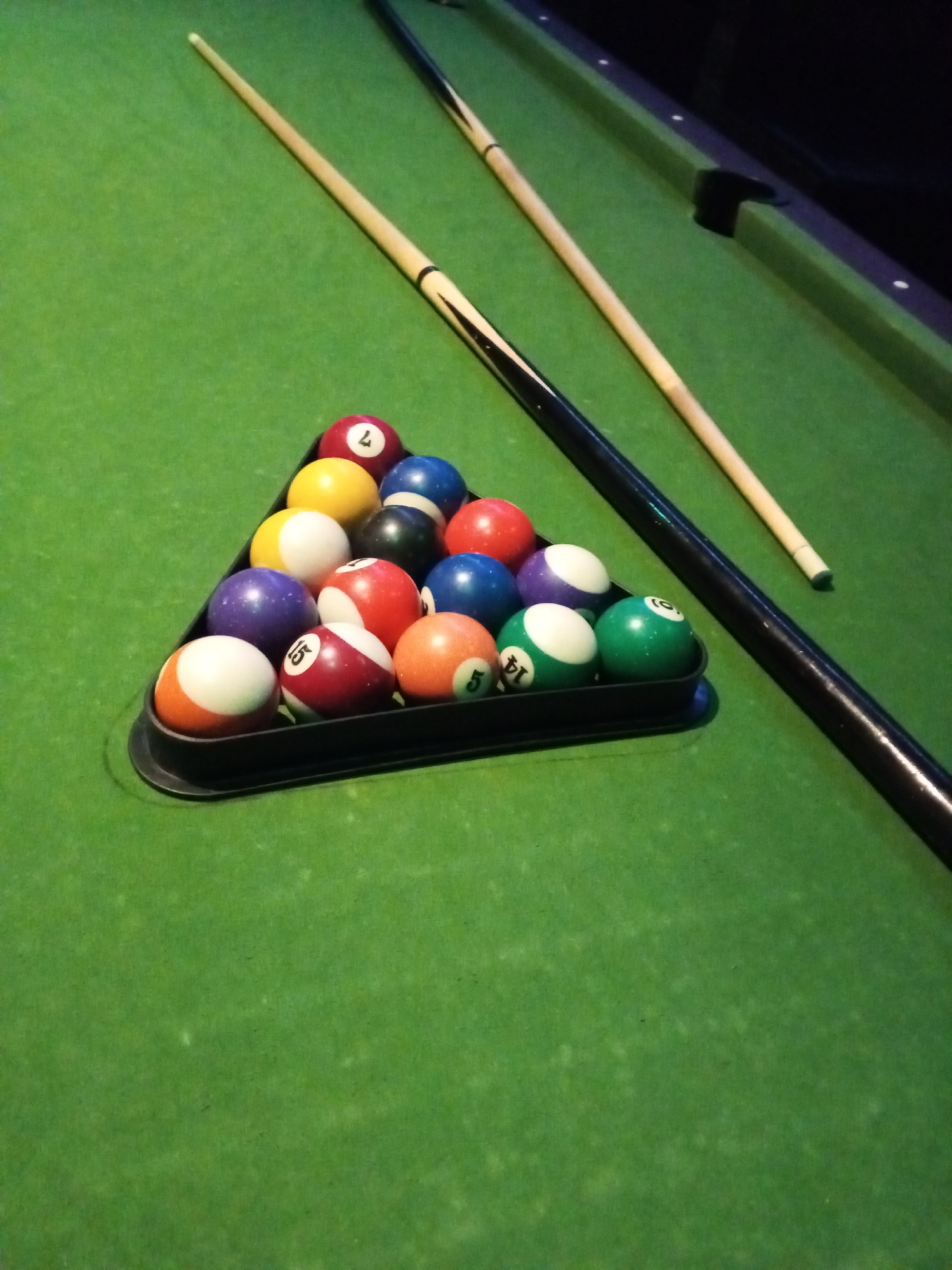 The image depicts a rack of pool balls, consisting of 15 numbered balls and one cue ball, arranged in a triangular formation on a green felt pool table. Two pool cues are also visible, resting on the table. One cue is positioned to the left of the rack, extending diagonally upwards, while the other is to the right, partially obscuring the rack and extending towards the bottom right corner. The balls are in various colors and numbers, and they appear to be standard pool balls. The environment is that of a typical pool hall or game room, with the green felt of the table being the dominant element in the foreground. The background is dark, suggesting an indoor setting with subdued lighting. There are no people visible in the image. The specific location of Lokoja, Nigeria, is not evident from the visual information presented in the image.