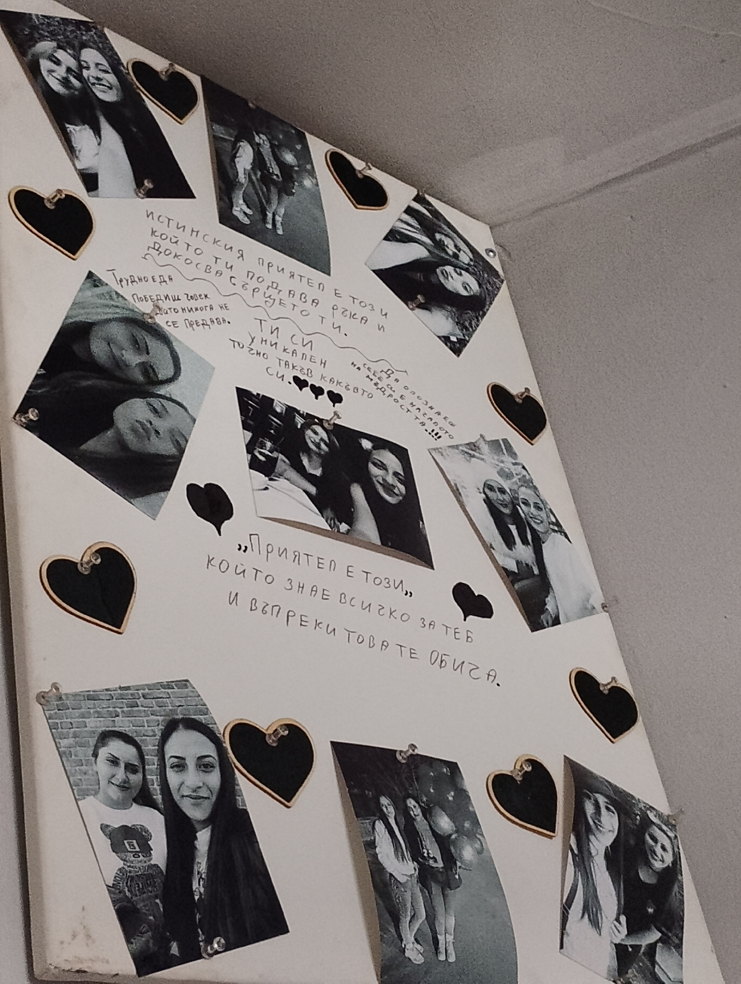 The image is a close-up, angled shot of a collage of black and white photographs and handwritten text on a white surface, likely a wall or board. The collage is decorated with several black heart-shaped embellishments.
The photographs depict groups of young women, primarily in casual settings, smiling and interacting with each other. Some photos show two women, while others show three or four. The handwritten text, in Bulgarian, appears to be messages or quotes. One prominent section reads: "ИСТИНСКИЯ ПРИЯТЕЛ Е ТОЗИ, КОЙТО ТИ ПОДДАВА РЪКА И ДОКОСВА СЪРЦЕТО ТИ." (A true friend is one who gives you a hand and touches your heart.) Another section reads: "ПРИЯТЕЛ Е ТОЗИ, КОЙТО ЗНАЕ ВСИЧКО ЗА ТЕБ И ВЪПРЕКИ ТОВАРЕ ОБИЧА." (A friend is one who knows everything about you and loves you anyway.)
The overall impression is a personal and sentimental display, possibly a memory board or a gift. The scene is static, with no discernible actions taking place beyond the display itself. The environment is indistinct, with a textured grey surface visible in the background, suggesting an indoor setting. There is no information in the image that indicates a specific location in Yambol, Bulgaria.