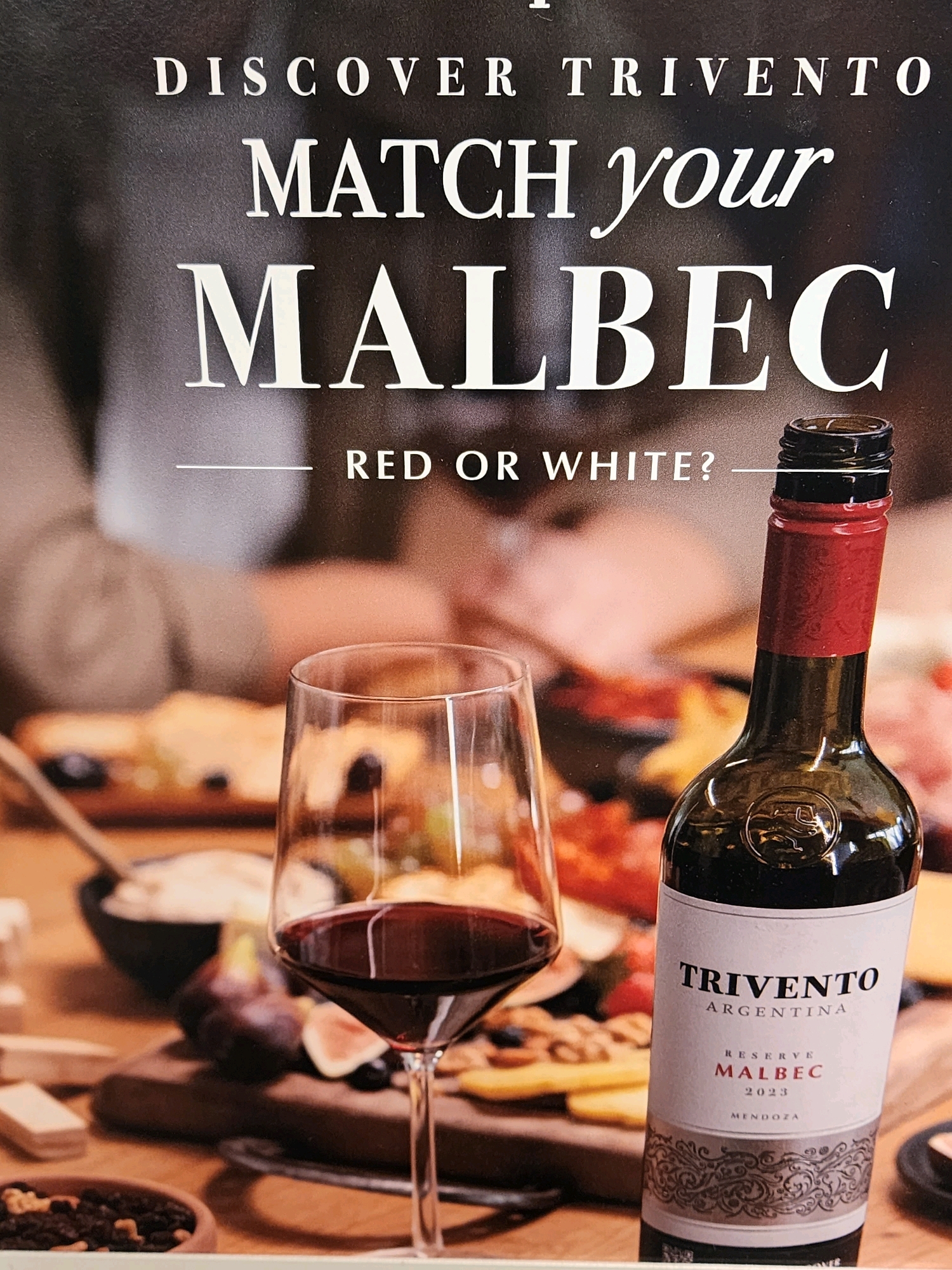 This is a close-up, eye-level shot of a wine bottle and a glass of red wine, placed on a table setting. The background is a blurred image of people at a dinner. The text on the bottle and in the foreground indicates that this is an advertisement for Trivento Malbec wine. The text at the top reads "DISCOVER TRIVENTO" and "MATCH your MALBEC". Below that, it asks "RED OR WHITE?". The wine bottle is dark green glass with a red label and a red foil cap. The label reads "TRIVENTO ARGENTINA RESERVE MALBEC 2023 MENDOZA". The glass is a clear wine glass filled with red wine. The table setting includes a cutting board with cheese, figs, and nuts. The overall scene suggests a social gathering centered around wine and food. The image appears to be a professional advertisement, likely for a winery or restaurant. The location is not specified in the image.