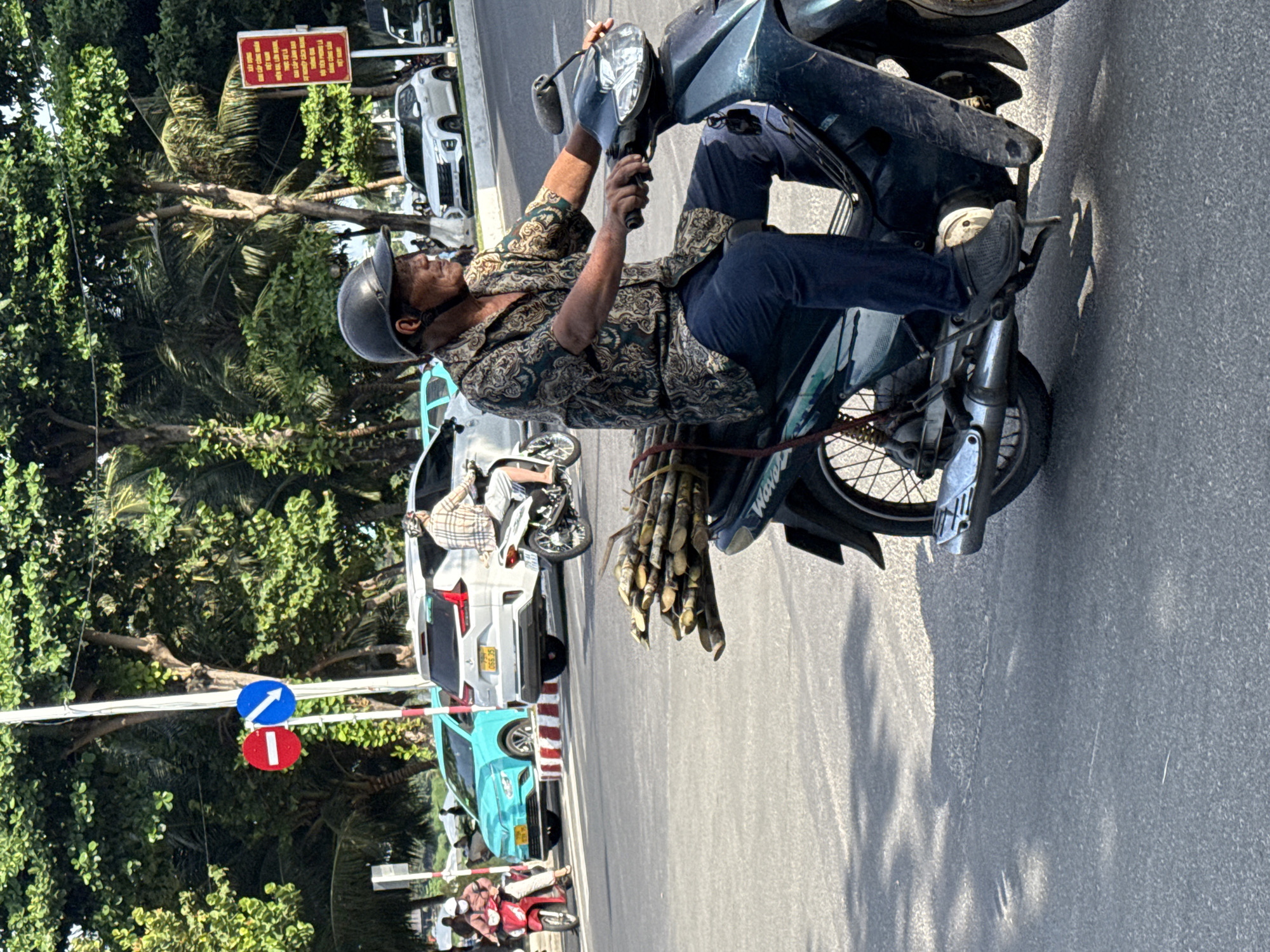 In Bac Nha Trang Ward, Vietnam, a middle-aged man wearing a helmet and a patterned shirt rides a motorcycle. He is carrying a bundle of sugarcane stalks tied to the back of his motorcycle. The man appears to be looking to his right and is holding a cigarette in his right hand. Other vehicles, including a turquoise car, a white SUV, and another motorcycle, are visible in the background, along with trees and street signs. The scene is on a paved road, suggesting a street environment in a Vietnamese city.