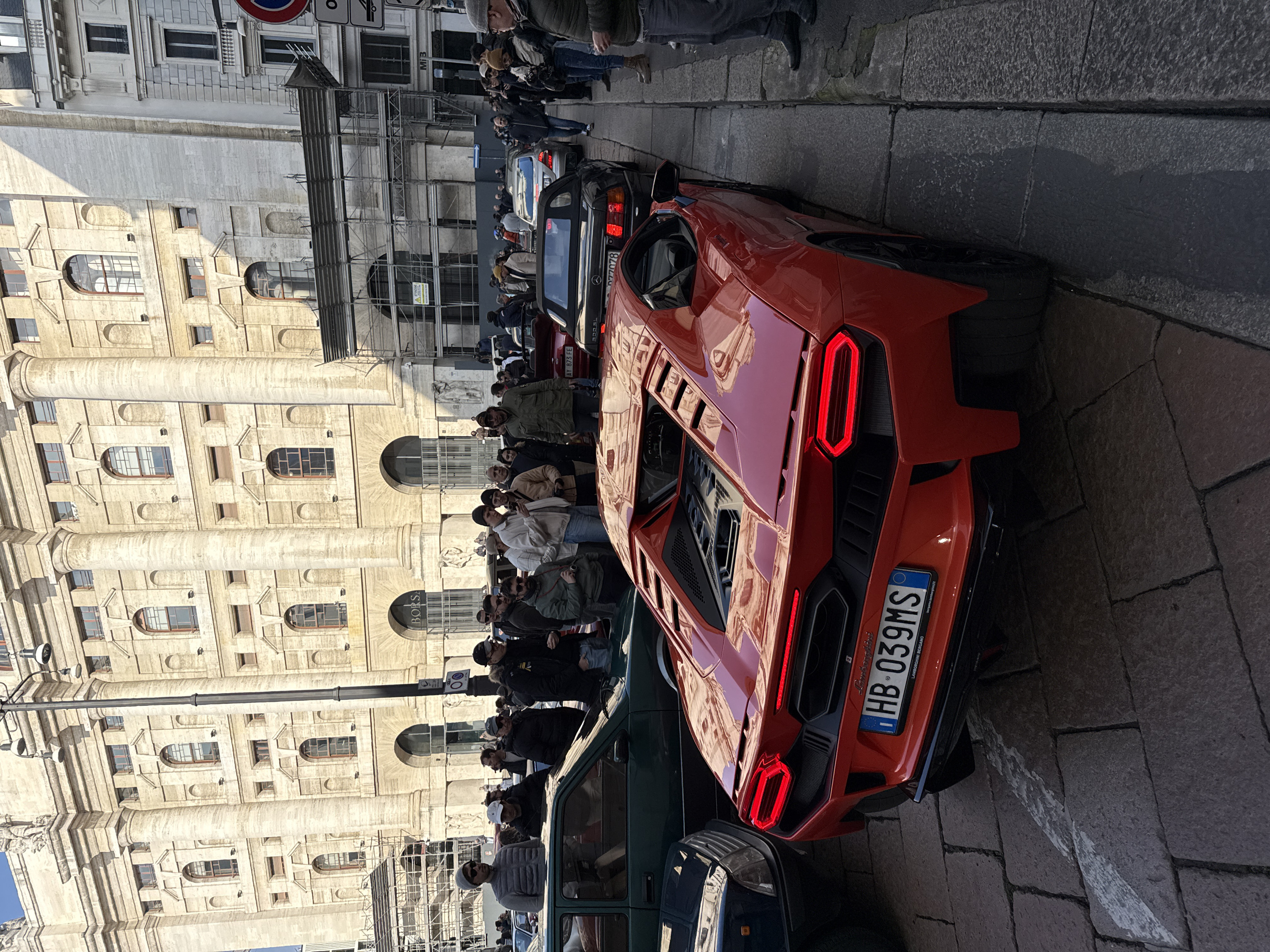 The scene captures a busy urban street in Milan, Italy, specifically Piazza Affari, with the imposing Palazzo Mezzanotte (Borsa Italiana building) in the background. A bright red Lamborghini Huracán STO, displaying Italian license plate "HB 039MS," is parked prominently on the cobblestone street, facing away from the viewer, with its rear engine bay and distinct LED taillights illuminated. To the left, a dark blue vehicle's front end and a dark green car are partially visible. Immediately behind the Lamborghini, a black Mercedes-Benz SL convertible is parked, showing a partial license plate "D77078." A crowd of approximately 30-50 individuals, predominantly men in various casual winter jackets and headwear, are gathered on the sidewalk and street, observing the parked vehicles. The multi-story Palazzo Mezzanotte is constructed of light-colored stone, featuring numerous arched and rectangular windows, large columns, and relief sculptures; scaffolding covers portions of its facade, indicating maintenance or construction. The street is paved with uneven cobblestones. The sky is clear and blue, and a black streetlight pole with multiple luminaires stands on the right, accompanied by various street signs.
