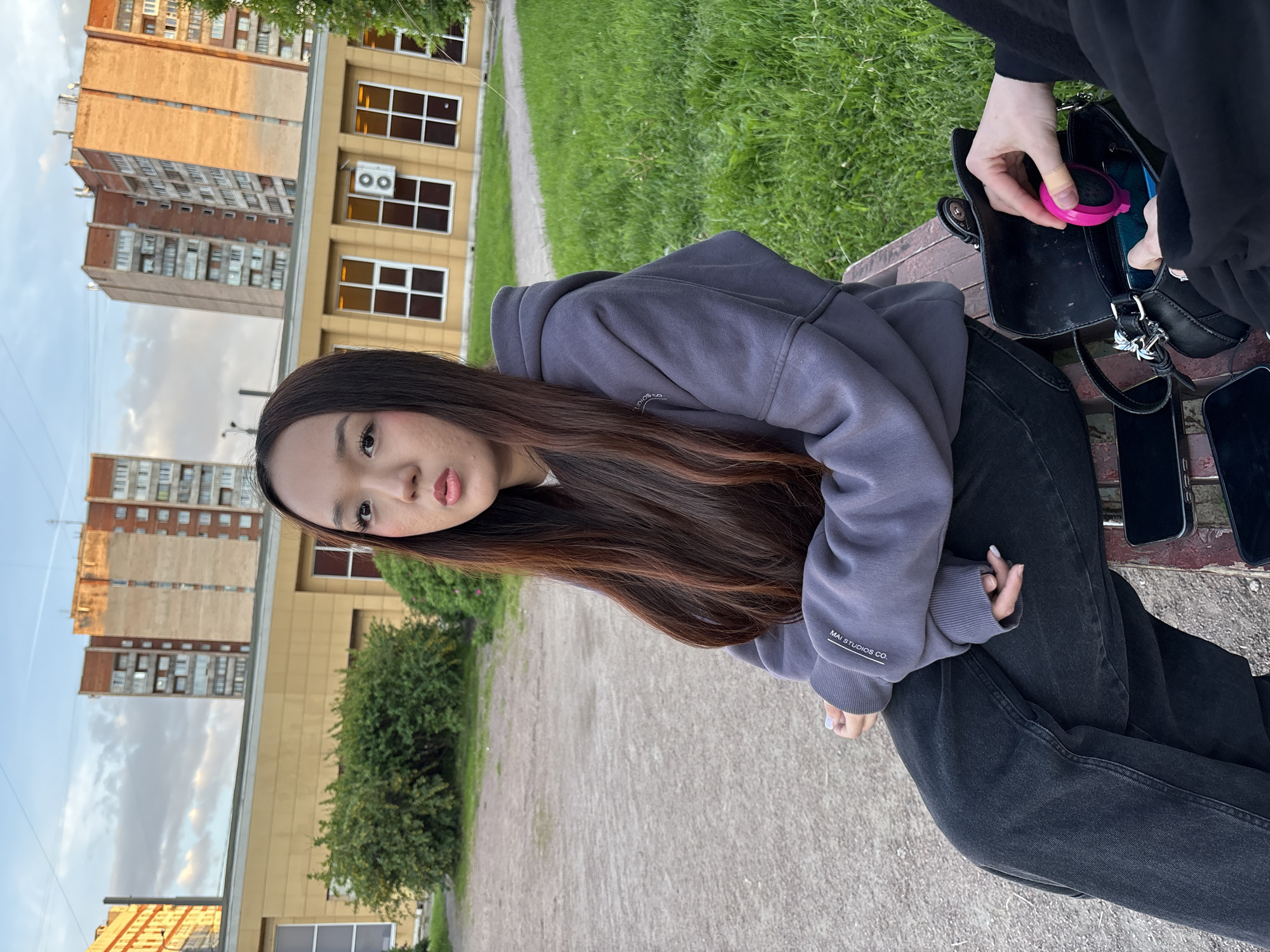 This media file captures a daylight scene in what appears to be an urban or suburban park or walkway in Saint Petersburg, Russia.
The primary subject is a young woman with long, dark hair, featuring reddish highlights, sitting on a wooden bench. She is wearing a grey/purple hooded sweatshirt, on which "MAI STUDIOS CO." is visible on the left sleeve, and black jeans. She is looking directly at the camera with a slightly pouting expression.
Next to her, another person's hands are visible, rummaging through a black handbag. This person is wearing black clothing. A small, bright pink, circular object, possibly a compact mirror or a portable speaker, is being held as it's either placed into or retrieved from the bag. Two smartphones are also resting on the bench beside the handbag.
The setting includes a paved path in the foreground and midground, flanked by grassy areas and low green bushes. In the background, several multi-story buildings are visible under a partly cloudy sky. One building is light yellow/brown with multiple rectangular windows and an air conditioning unit on its facade. Beyond this, a taller, multi-storied apartment building, made of light brown concrete, stands prominently, characteristic of Eastern European architecture. The lighting suggests it is daytime, possibly late afternoon, with clear weather.