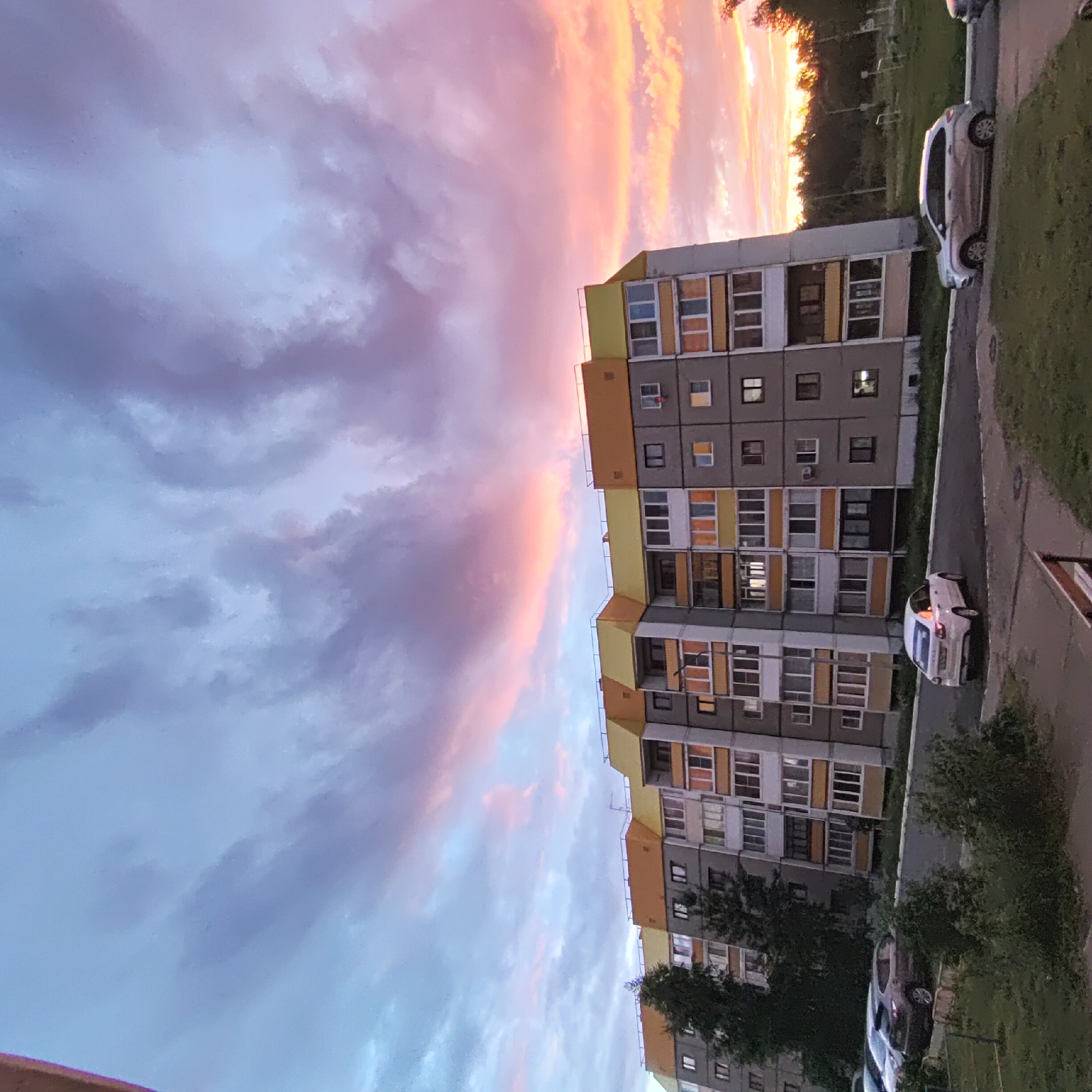 The image depicts a residential building in the city of Markovskiy, Russia, under a dramatic sunset sky. The building is a multi-story apartment complex, characterized by its grey facade and colorful yellow and orange accents on the upper floors and roofline. Numerous windows and balconies are visible, some with lights on, suggesting occupancy. In the foreground, parked cars are visible on the asphalt road and in a grassy area. A white sedan is in motion on the road, while a silver SUV is parked. The sky is filled with swirling clouds in shades of blue, purple, pink, and orange, indicative of a sunset. Trees and foliage are present on the ground and behind the building, with a denser treeline visible in the distance. The overall scene captures a common urban landscape during a picturesque sunset.