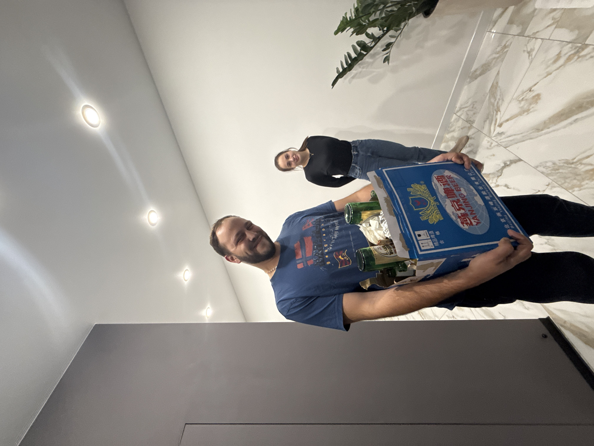Two individuals are in a brightly lit interior space. A man in the foreground is carrying a case of Yanjing Beer. The woman in the background is smiling. The setting includes a white marble floor and a plant in a pot. There are recessed lights in the ceiling. The location is unknown and cannot be ascertained from the image.