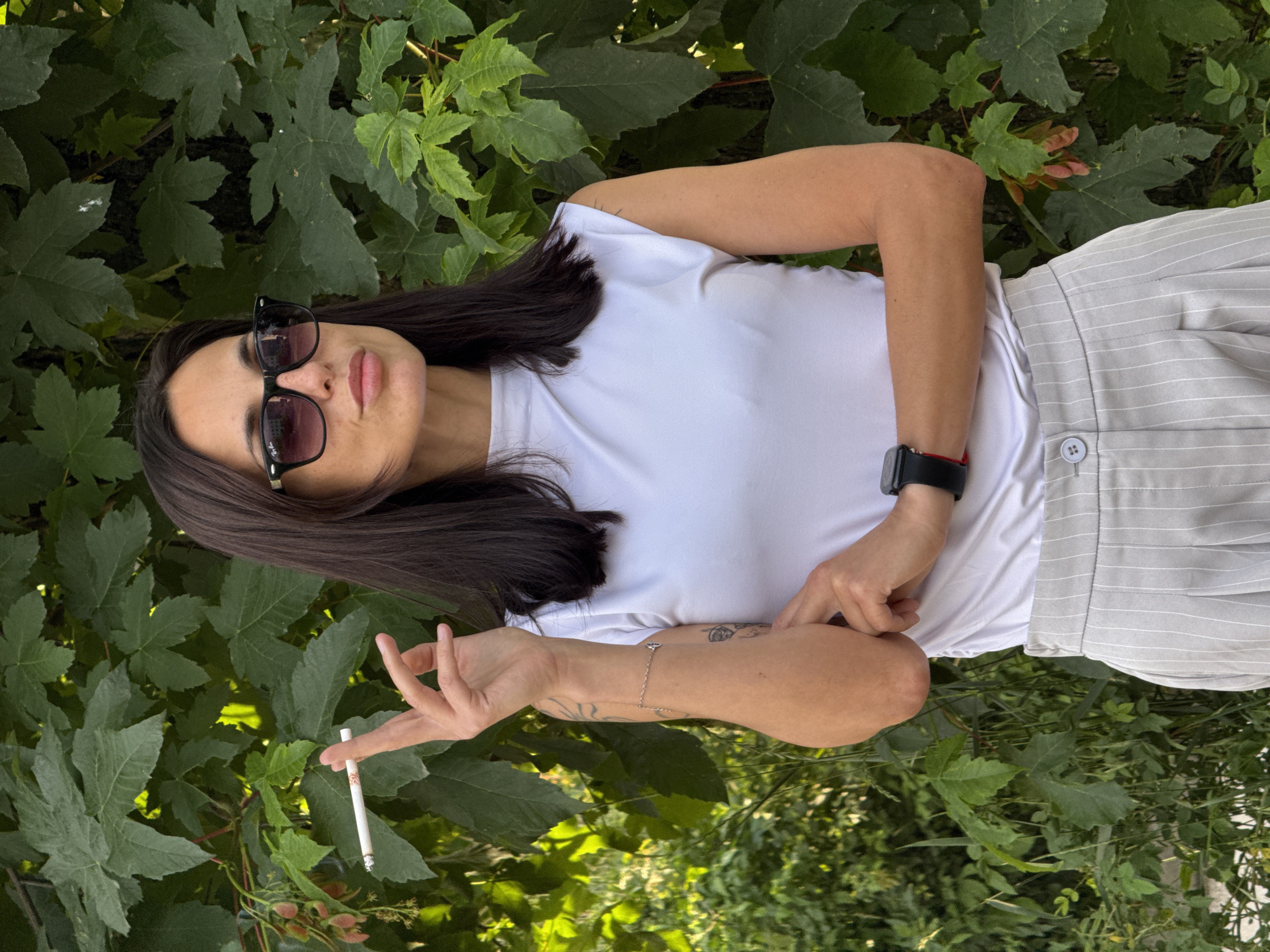 A young woman with dark hair and sunglasses is posing outdoors. She is wearing a white t-shirt and striped grey pants, and she has a watch on her left wrist. In her left hand, she holds a cigarette. The background consists of green leaves and foliage. The location is not specified, however the request stated that the location is in the city of Pleven, Bulgaria.