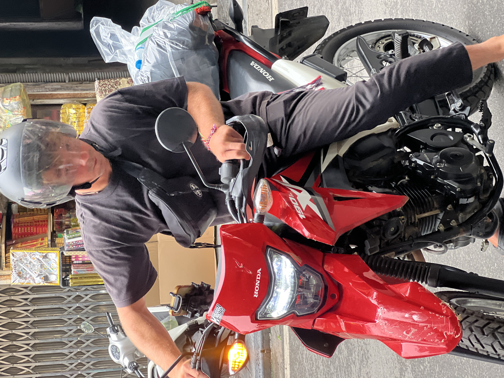A young man wearing a black t-shirt and black pants sits on a red and white Honda 190L motorcycle. He wears a silver helmet with a clear visor. A black fanny pack is worn across his chest, and a large plastic bag filled with items is secured to the back of the motorcycle. The man's right hand is on the motorcycle's handlebar. He appears to be in an urban environment, possibly a street in Hanoi, Vietnam, with a storefront and a metal gate visible in the background. The motorcycle's headlight is on, and its front fender shows signs of damage.