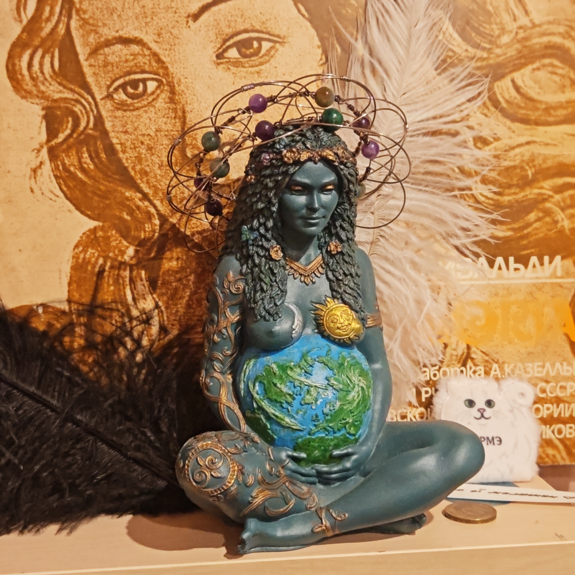 A teal-colored statue of a pregnant woman, representing a Mother Earth figure, is the central subject of this indoor scene. The statue is seated with crossed legs, her hands gently resting on her belly, which is depicted as a detailed globe with blue oceans and green landmasses. Her skin is a deep teal, and her long, curly hair, which appears to be made of vines or leaves, is accented with gold. A wire halo or crown adorned with purple and green beads encircles her head. She wears a golden necklace, and golden ornaments decorate her arms and legs. A golden sun emblem is on her right breast, and a crescent moon on her left. Her expression is serene and peaceful.
The statue rests on a light-colored surface, possibly a shelf or table. In the background, a large, sepia-toned illustration of a woman's face, with a prominent eye and wavy hair, is visible on a vertical surface. A large white feather is positioned to the statue's right, and a large black feather lies on the surface to its left. To the right of the statue, a small, fluffy white cat plushie with green eyes and a pink nose is present, and a coin lies on the surface in front of it.
Visible text in Cyrillic is seen on the background material: "БАЛЬДАЙ" is partially visible at the top right. Below it, "аботка А. КАЗЕМЛЬ," "СССР," and partially "ОРИЙ" are discernible. The letters "РМЭ" are printed on the white cat plushie. The overall setting is a curated display, possibly in a home or spiritual space in Novo-Nikol'skoe, Russia, with an emphasis on symbolism related to nature, creation, and femininity.