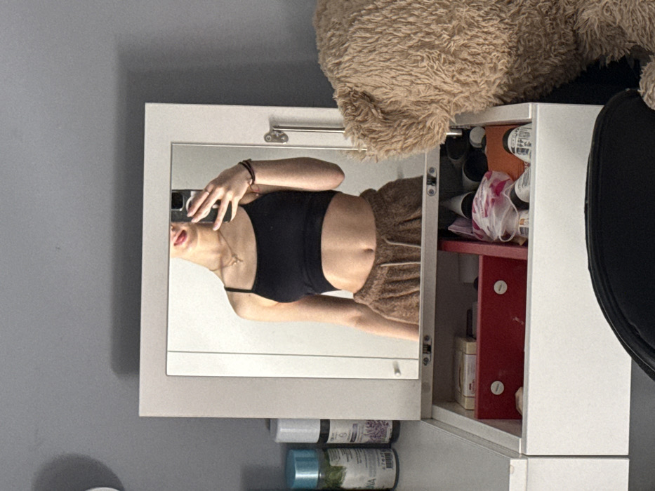 The image depicts an interior scene within a residence located in Sofia, Bulgaria. A subject, appearing to be a young female, is captured in a mirror selfie, partially obscuring her face with a dark-colored smartphone held in her right hand. She is wearing a black spaghetti-strap crop top, exposing her midriff, and light brown, textured (possibly fuzzy) lounge pants or sweatpants. A golden-colored necklace with a small pendant is visible around her neck, and a dark bracelet is on her left wrist.
The mirror is framed in white and mounted on a white vanity or dressing table. To the left of the mirror, on the table surface, are several upright bottles, including a light blue-capped green bottle and a white-capped bottle labeled with lavender imagery. Below the mirror, an open drawer or compartment reveals organized contents: a red compartmentalized tray containing small items like white containers or boxes, and to its right, various toiletries, cosmetics, and small plastic bags.
To the far right of the image, a large, light-brown, fluffy teddy bear is partially visible. In the bottom foreground, the dark backrest of a chair is present. The wall behind the vanity and to the left of the mirror is a plain grey, with a circular white object (possibly a clock face) partially visible in the upper left corner. The reflection in the mirror shows a white wall or door behind the subject.