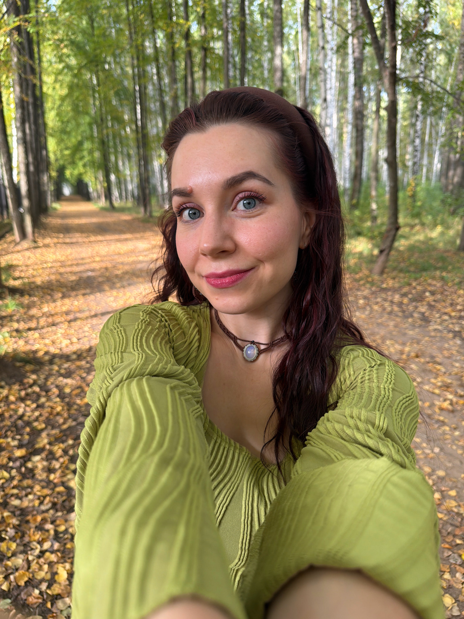 A medium shot captures a young woman from the chest up, standing in a tree-lined park in Chaikovsky, Russia. She has long, dark, wavy hair, a light complexion, and blue eyes. She is wearing a lime green textured top and a brown necklace with a circular pendant. The background shows a path lined with trees, with fallen yellow leaves scattered on the ground, indicating an autumn setting. Sunlight filters through the trees, creating a dappled effect.