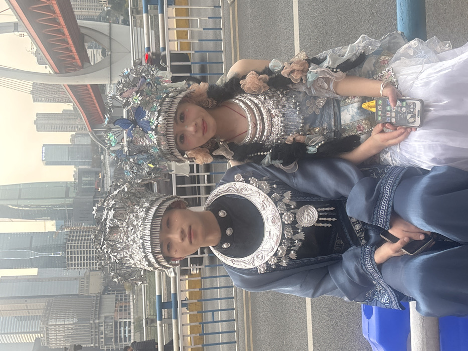 Two young East Asian individuals are seated outdoors on an elevated pathway or bridge in Yuzhong District, Chongqing, China. They are positioned in front of a metal railing, with a road behind them and a dense urban skyline forming the backdrop.
The male on the left wears a dark blue traditional-style robe with silver embroidery, a black undergarment, and an elaborate silver-colored necklace featuring a large circular pendant and dangling pieces. His head is adorned with a multi-tiered silver headpiece detailed with metallic leaf patterns. He holds a dark-colored smartphone in his lap.
The female on the right wears a light blue and grey floral-patterned sleeveless top over a white pleated skirt. She has multiple silver-colored necklaces, one with a red cord, and her dark braided hair is embellished with light pink and blue fabric flowers. Her silver-colored headpiece features prominent blue butterflies and smaller floral elements. She holds a white-cased smartphone with a yellow charging cable connected, displaying app icons.
Both individuals are looking forward with neutral to slightly smiling expressions.
The background features numerous modern skyscrapers under an overcast sky, characteristic of Chongqing's urban landscape. A large red-orange suspension bridge with white concrete tower supports is visible to the right, spanning a body of water. Blurred vehicles are present on the road behind the individuals and the railing.