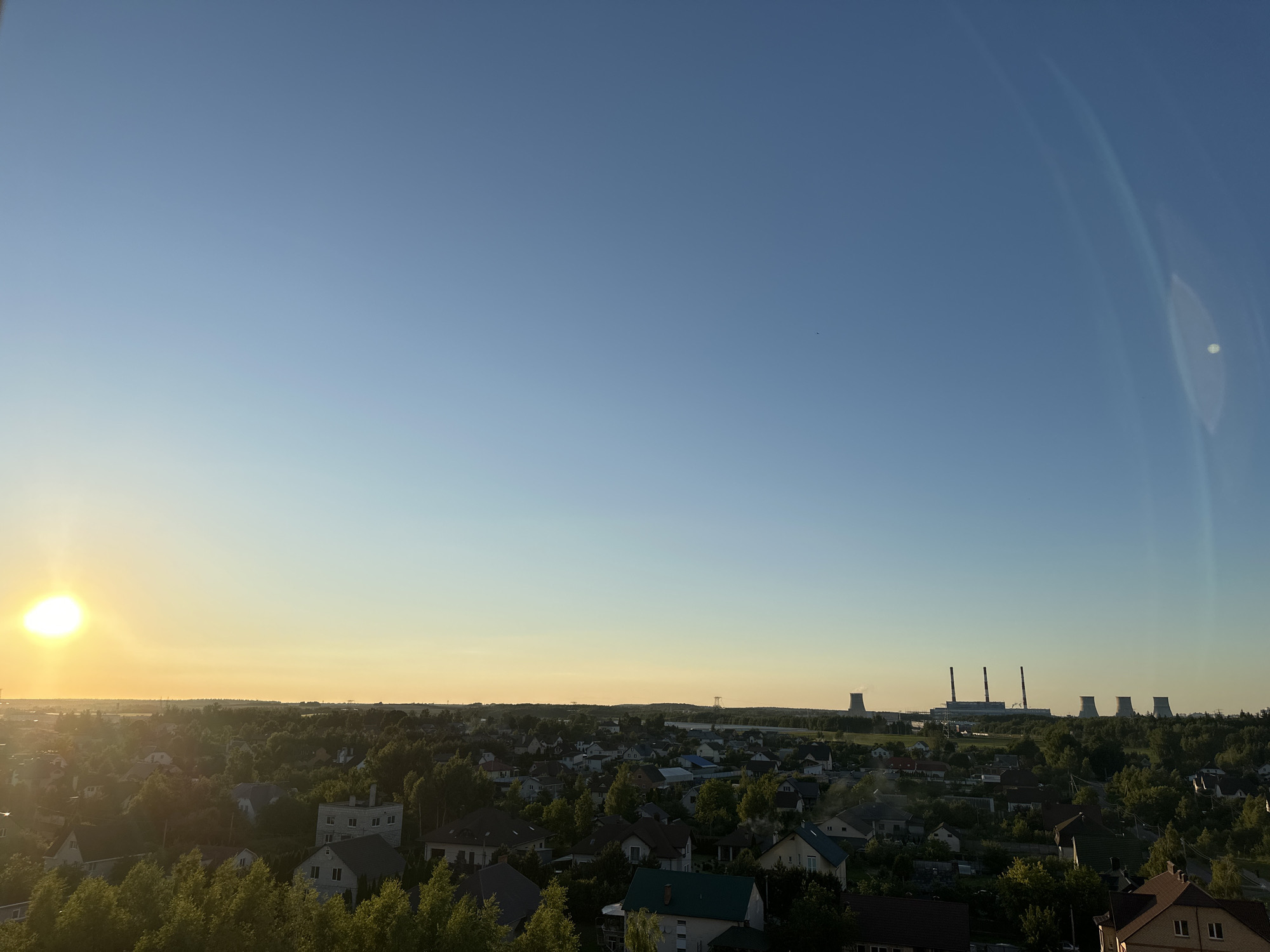 The image captures a panoramic view of a suburban or rural landscape in or near Шчомысліцкі сельскі Савет, Belarus, during a sunset. The sun, a bright orb radiating warm light, is positioned on the left side of the frame, casting a golden glow across the horizon. The sky is a clear, gradient blue, transitioning to yellow and orange near the horizon.
In the midground, a dense collection of residential buildings, likely houses with pitched roofs and varying architectural styles, are nestled among abundant green trees. The foliage is lush, suggesting a temperate climate and possibly the warmer months.
Further in the distance, a prominent industrial complex with several tall, imposing cooling towers is visible. These structures, characteristic of power plants or similar facilities, stand out against the skyline and the surrounding greenery. The overall scene conveys a sense of a community situated between natural landscapes and industrial infrastructure, bathed in the soft light of the setting sun.
