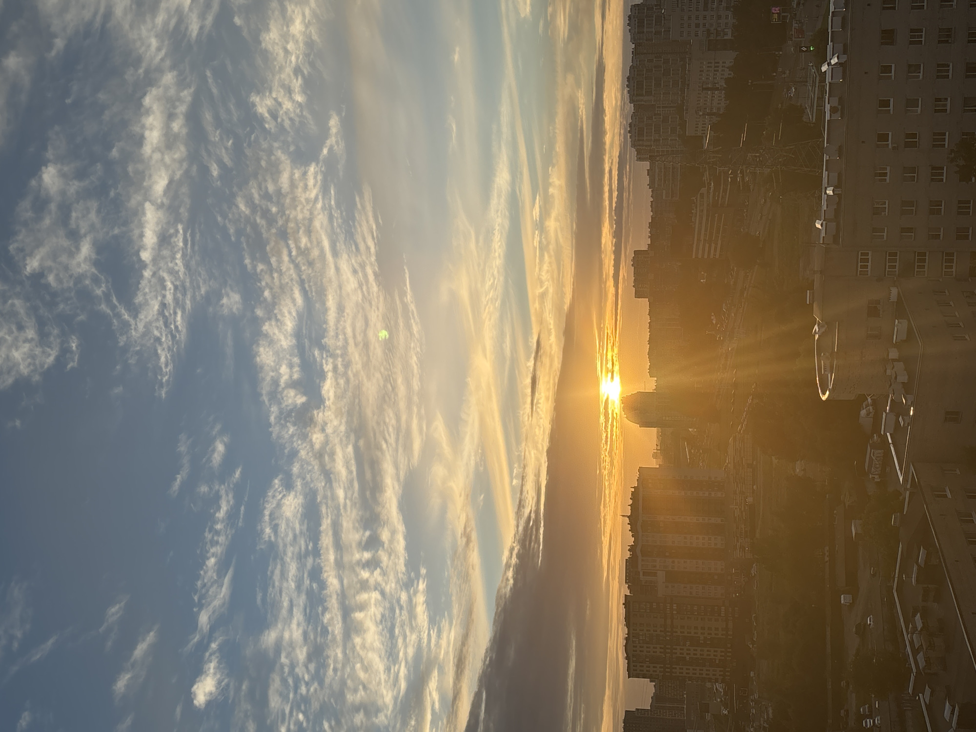 This media file is a landscape photograph taken vertically, capturing a sunset over a city. The sky is filled with wispy, streaky clouds, primarily in shades of white and light yellow, with a gradient towards a soft blue. The sun is a bright, glowing orb positioned on the right side of the image, low in the sky, casting long rays of golden light that illuminate the clouds and buildings.
The scene depicts an urban environment with numerous tall apartment buildings. These buildings are mostly rectangular in shape and appear to be densely packed together. Some buildings have visible windows, and a few have rooftop structures like air conditioning units. The warm, golden light of the sunset casts a long shadow over the city, creating a silhouette effect in some areas. The overall atmosphere is serene and peaceful, evoked by the soft light and the quiet grandeur of the cityscape at dusk. The time of day is clearly sunset, indicated by the low angle of the sun and the warm, diffused lighting. No people or specific objects are discernible as the main subjects, with the focus being on the interplay of light, clouds, and architecture. There is no visible text in the image.