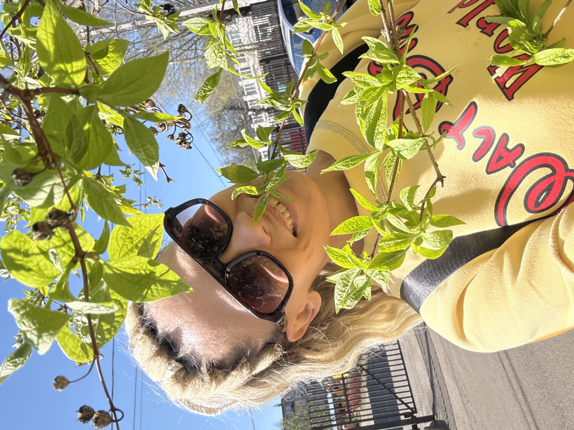 A woman wearing a yellow sweatshirt with red lettering and sunglasses is smiling as she poses outdoors. She is positioned among branches of a tree with vibrant green leaves. Her blonde hair is visible, and she has a fair complexion. The background includes buildings, a vehicle reflection in her glasses, a fence, and a clear blue sky. The photo appears to have been taken in Rusanovo, Russia.