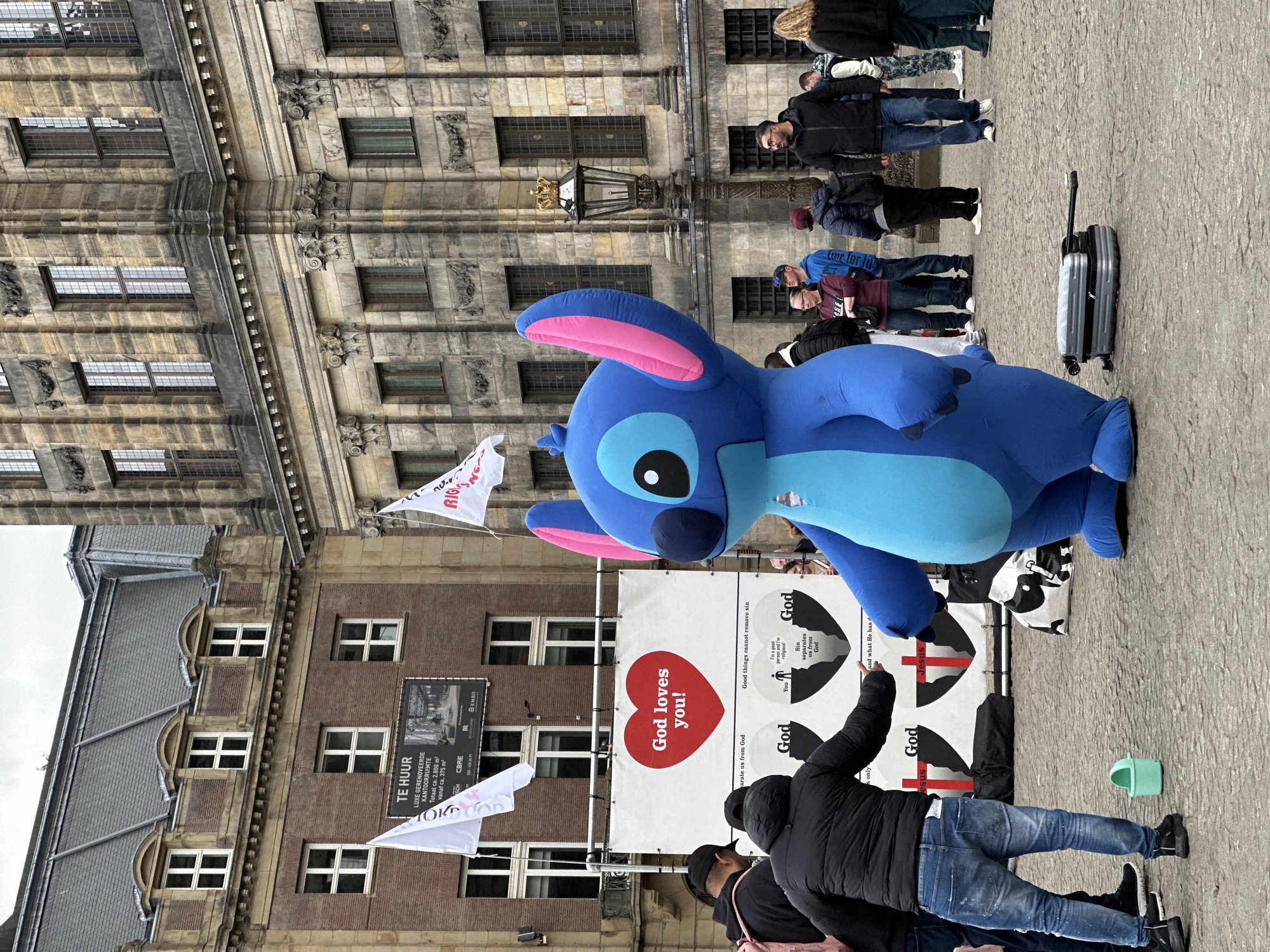 The image shows a public space in Amsterdam, Netherlands. A large, blue inflatable Stitch character stands in front of a banner that reads "God loves you!" with illustrations about sin and religion. A man in a black jacket points to the banner, engaging with its message. A group of people stands in a line in the background, some with luggage. The backdrop includes a historic building with windows and a "Te Huur" sign, suggesting property for rent. Flags are attached to the banner.