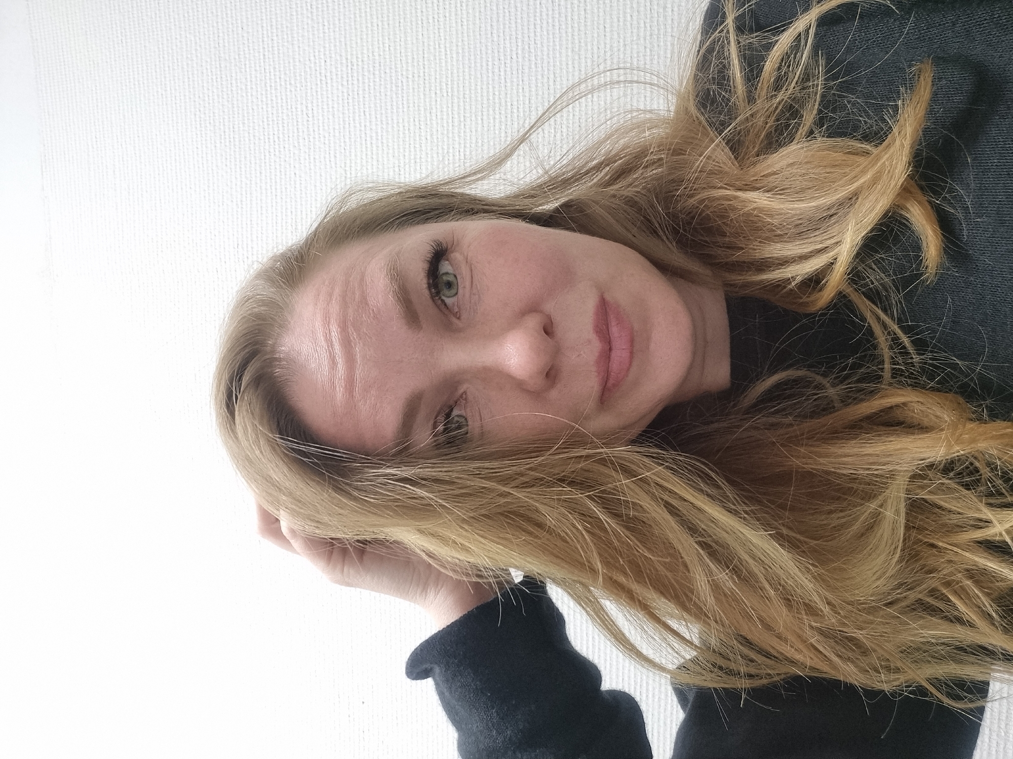 A woman with long, blonde hair is pictured close-up against a white wall. She is wearing a dark-colored, long-sleeved garment. Her face is slightly tilted, and she appears to be resting her head on her hand. Her eyes are light-colored, and she has fair skin. No specific details connect the image to Kemul, Russia, or provide contextual information about the scene or individuals involved.