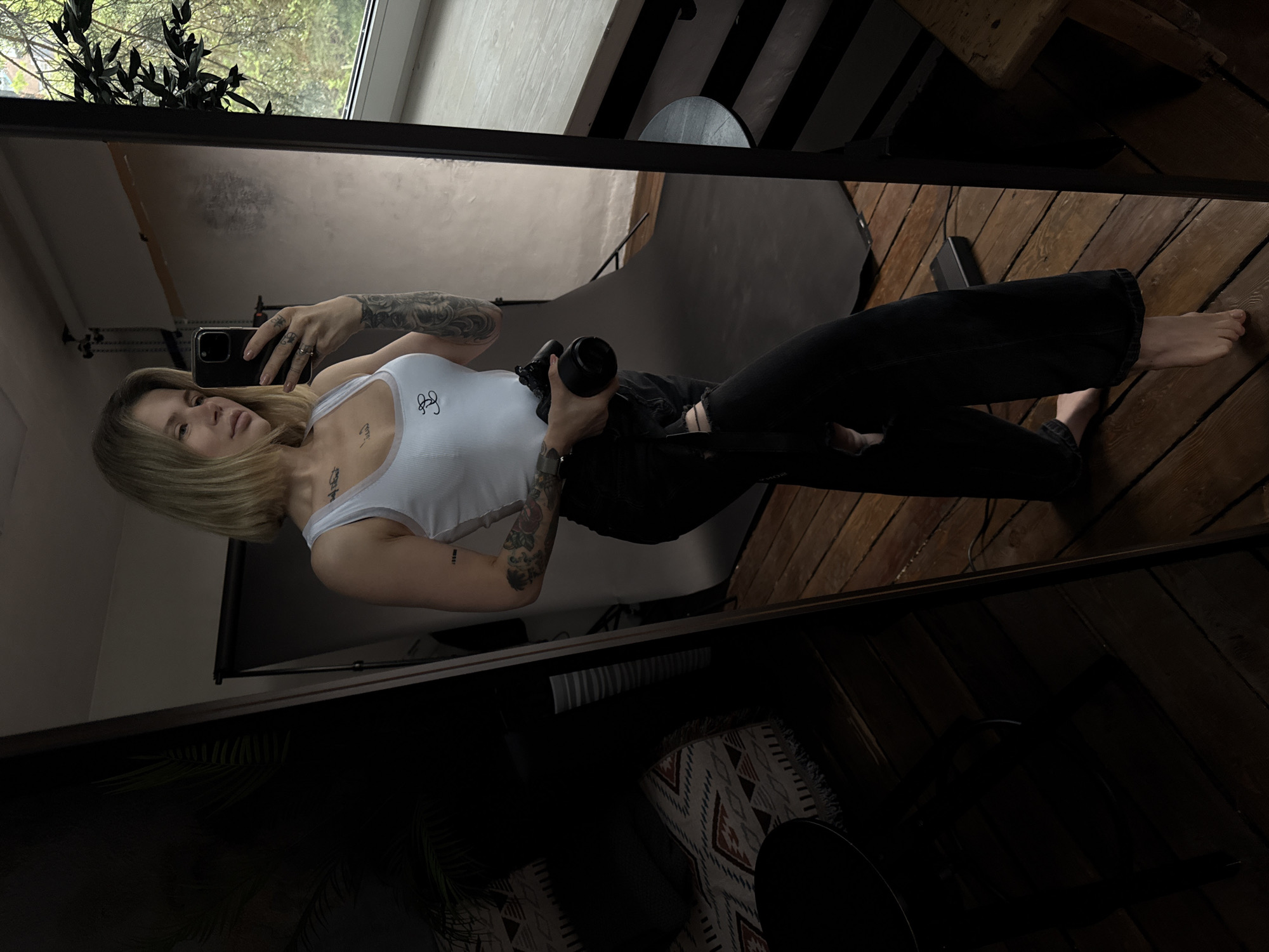 A young woman with blonde hair, wearing a white tank top and black jeans, stands barefoot in a room. She holds a camera in her right hand and a phone in her left, appearing to take a selfie in a mirror. The room contains a backdrop, suggesting a photography setup. The floor is wooden. The image does not contain sufficient information to determine if it was taken in Gorno-Altaysk, Russia.