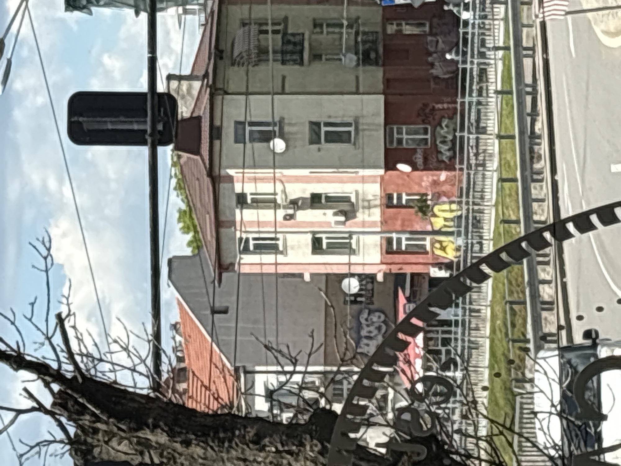 The image depicts a street scene in an urban area of Unknown, Unknown, viewed from a slightly low angle looking up. In the foreground, there are bare tree branches and an iron clock. Beyond the clock, a multi-story building with a facade divided into sections of beige, red, and gray with visible windows and some graffiti. A street with greenery alongside it runs adjacent to the building. A traffic light is visible to the left, positioned against a backdrop of a cloudy sky. The scene appears to be taken during daylight hours, showcasing a typical urban landscape.