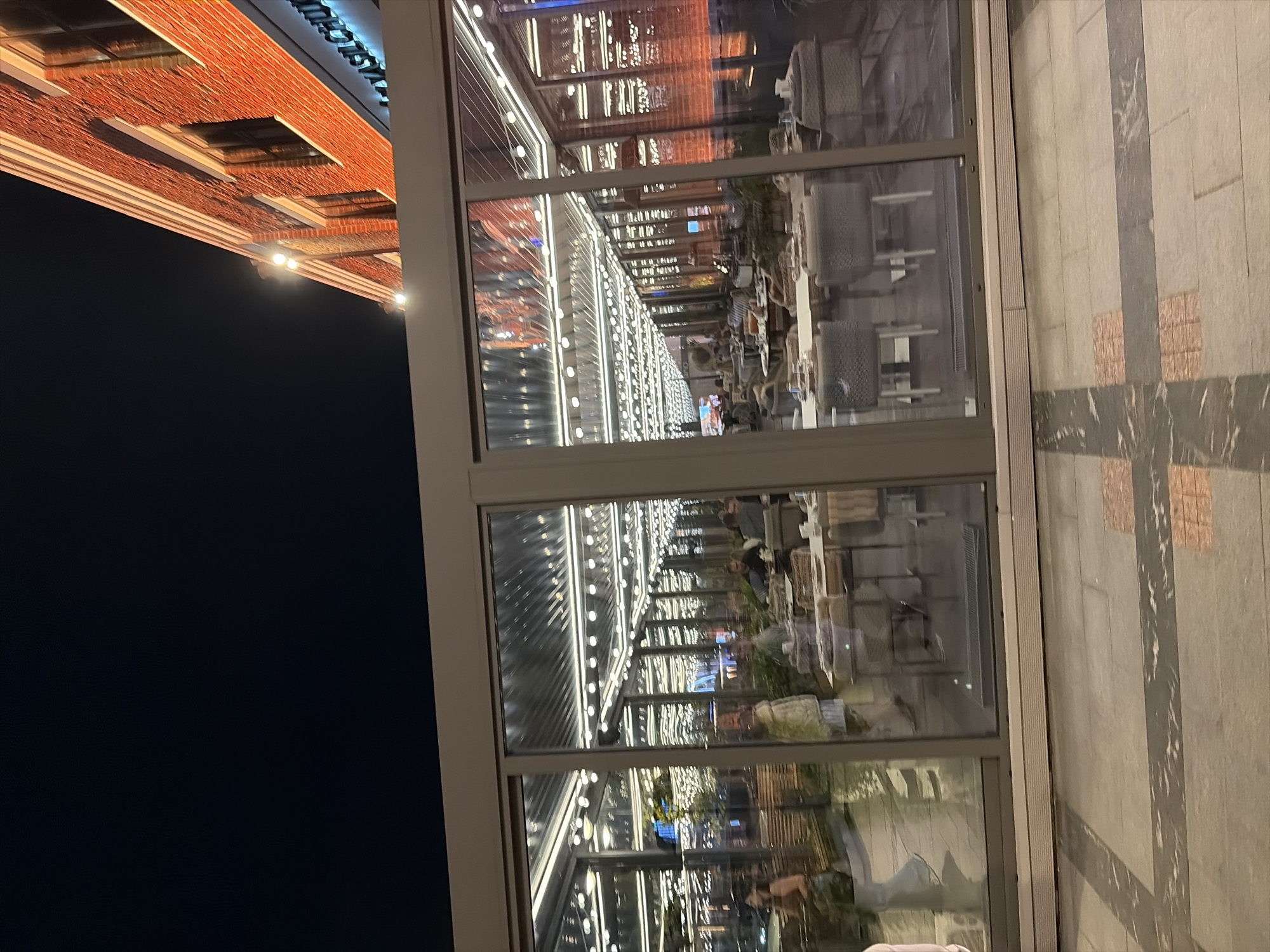 The image depicts a nighttime scene of a modern restaurant with an outdoor seating area enclosed by glass walls. The restaurant is situated next to a red brick building with large windows and is illuminated by strings of fairy lights overhead. Inside the restaurant, several tables are set with chairs, and people are seated, engaged in dining and conversation. The foreground shows a paved walkway with a decorative pattern. Based on the architectural style of the building and the presence of the text "AYASICI CHU" visible on the building facade, the location is likely in Moscow, Russia.