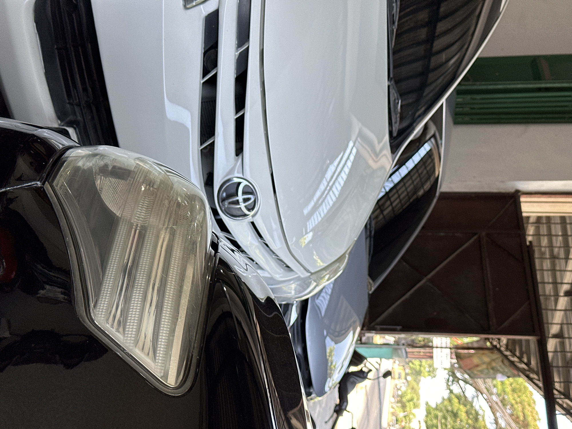 In Bulakrejo, Indonesia, a close-up shot captures the front of three cars parked under an open-air garage. The vehicle in the foreground is black, with its right headlight clearly visible and showing signs of wear. Behind it, a white Toyota is parked, its grille and emblem in sharp focus. The third car, a light gray sedan, is visible behind the white Toyota. The background reveals a street scene with trees, a motorcycle, and buildings, suggesting an urban setting. The lighting is natural, indicating it is daytime.